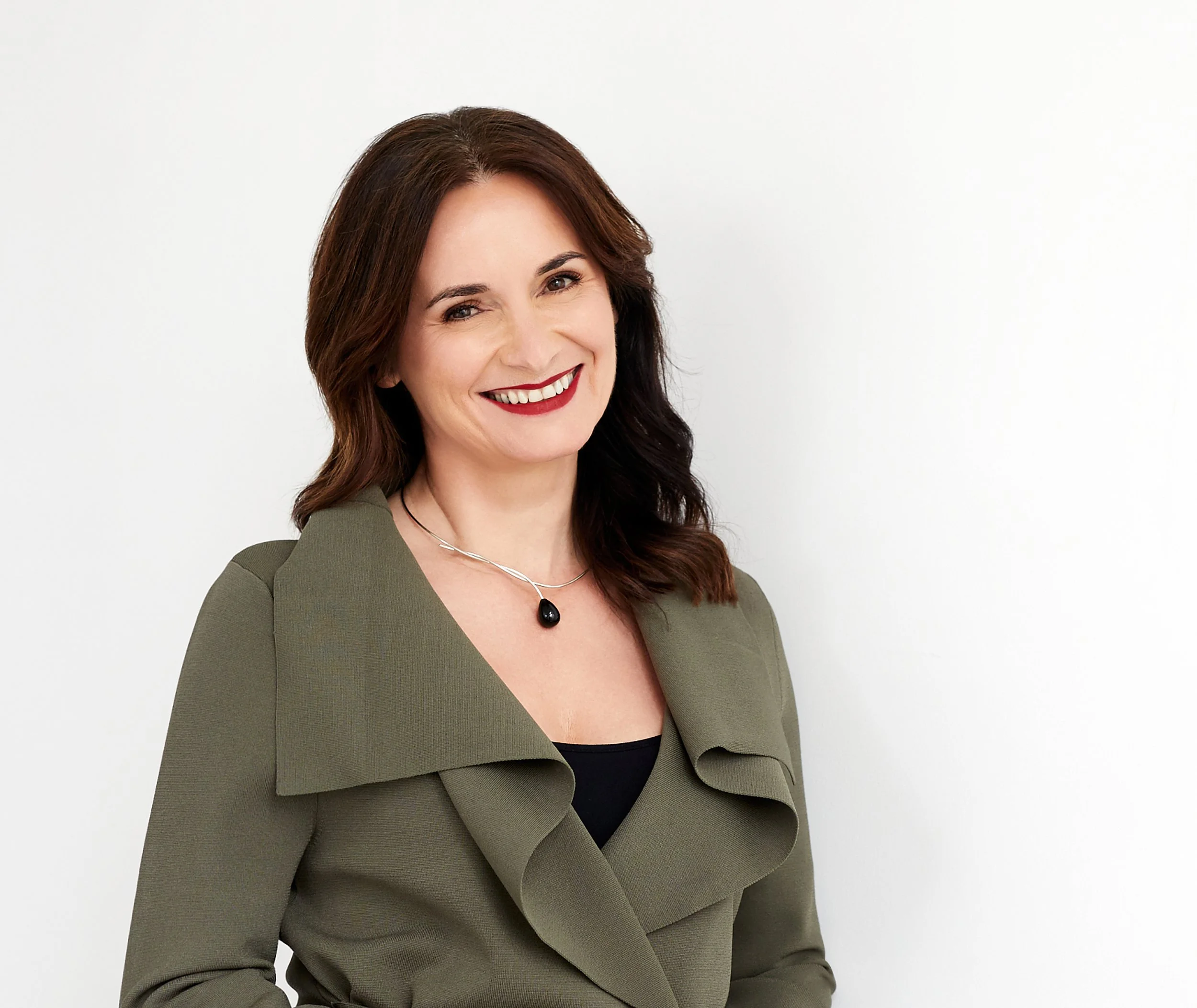 Photo of Zora Artis, smiling at the camera. She has dark long wavy hair, wears a dark-coloured blazer over a black shirt.