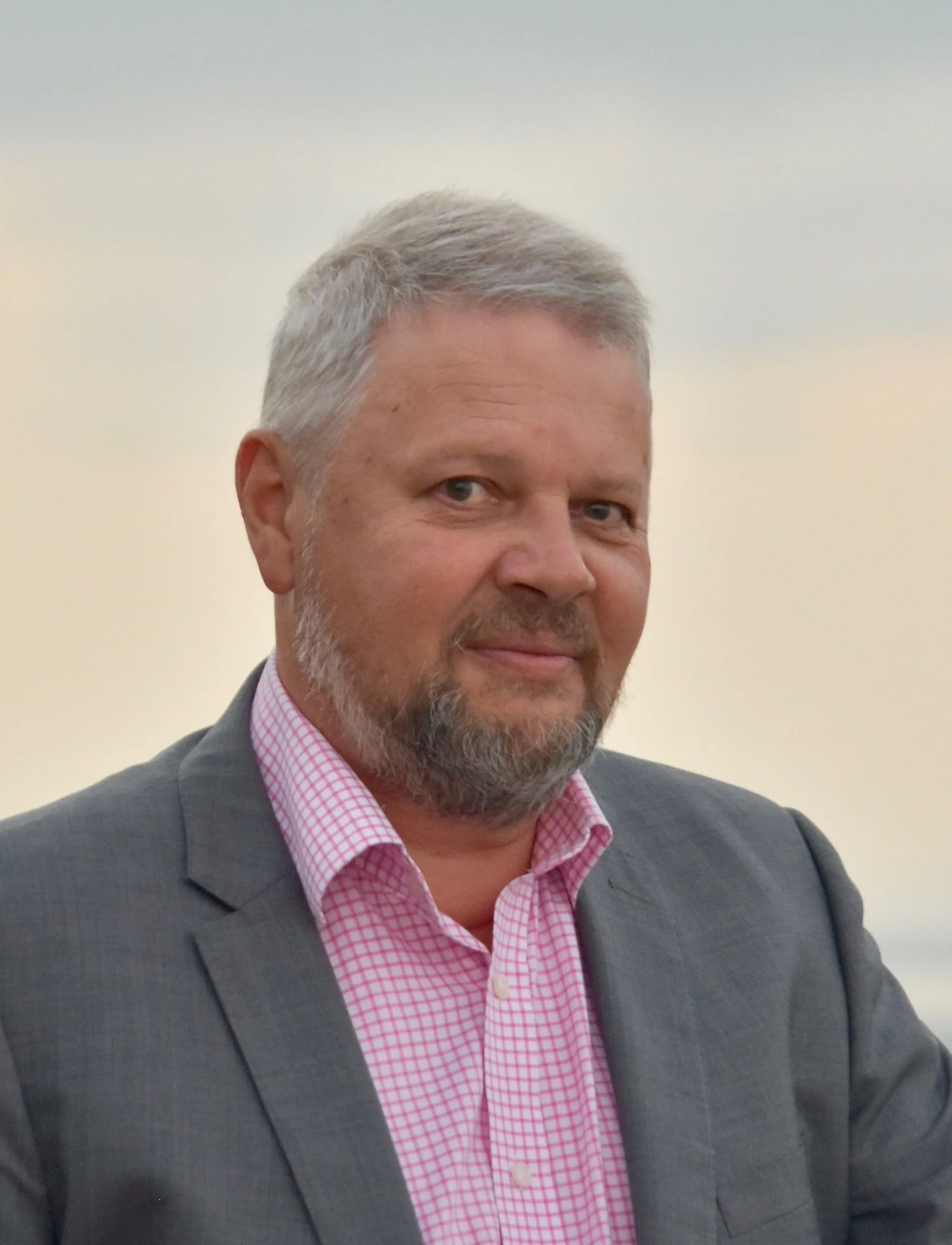 Photo of Wayne smiling at the camera. He has short grey hair, beard and moustache. He wears a grey blazer over a pink check shirt.