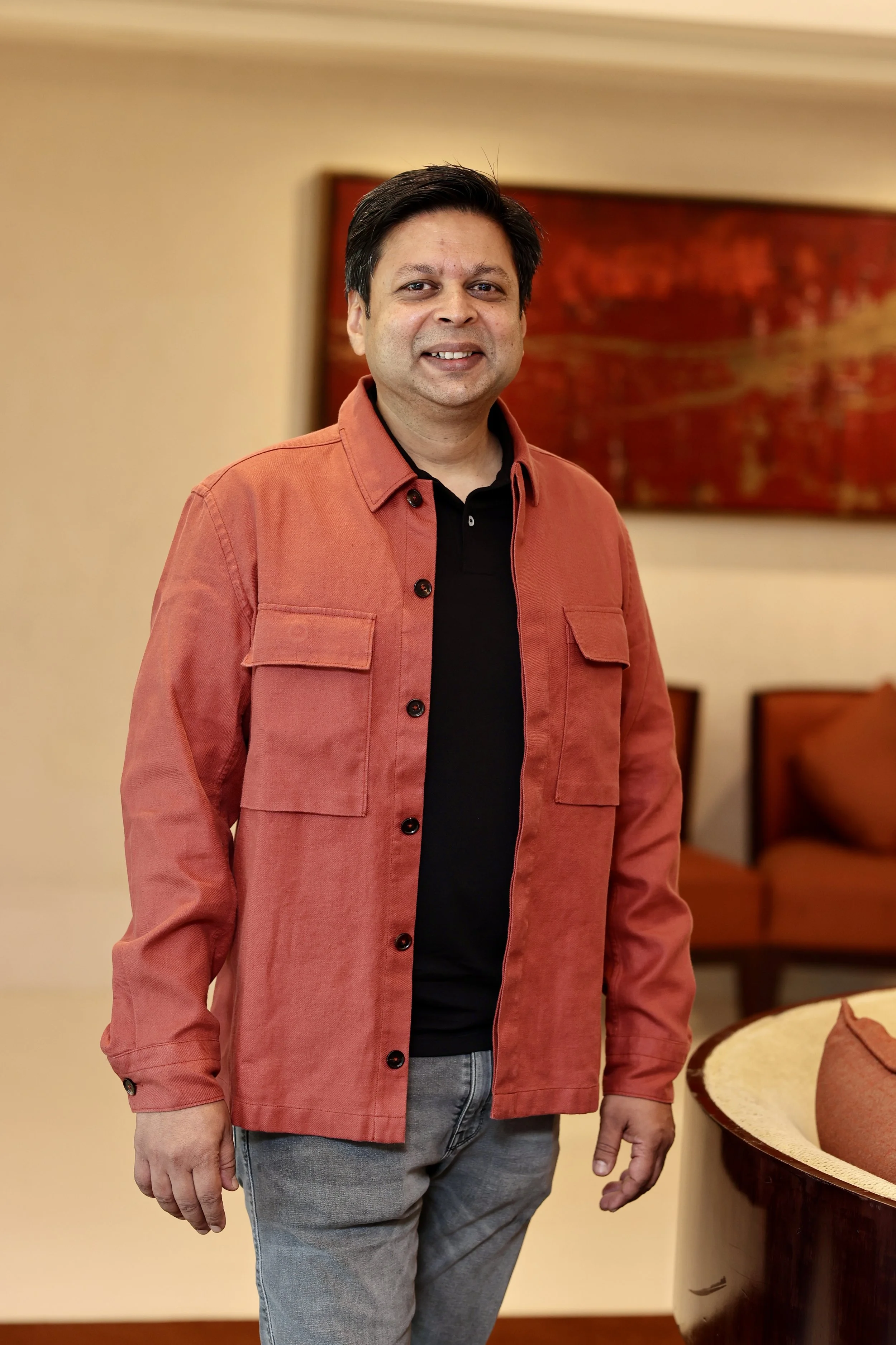 Photo of Anshuman smiling at the camera. He has short dark hair, and wears a red coloured jacket over a black shirt.