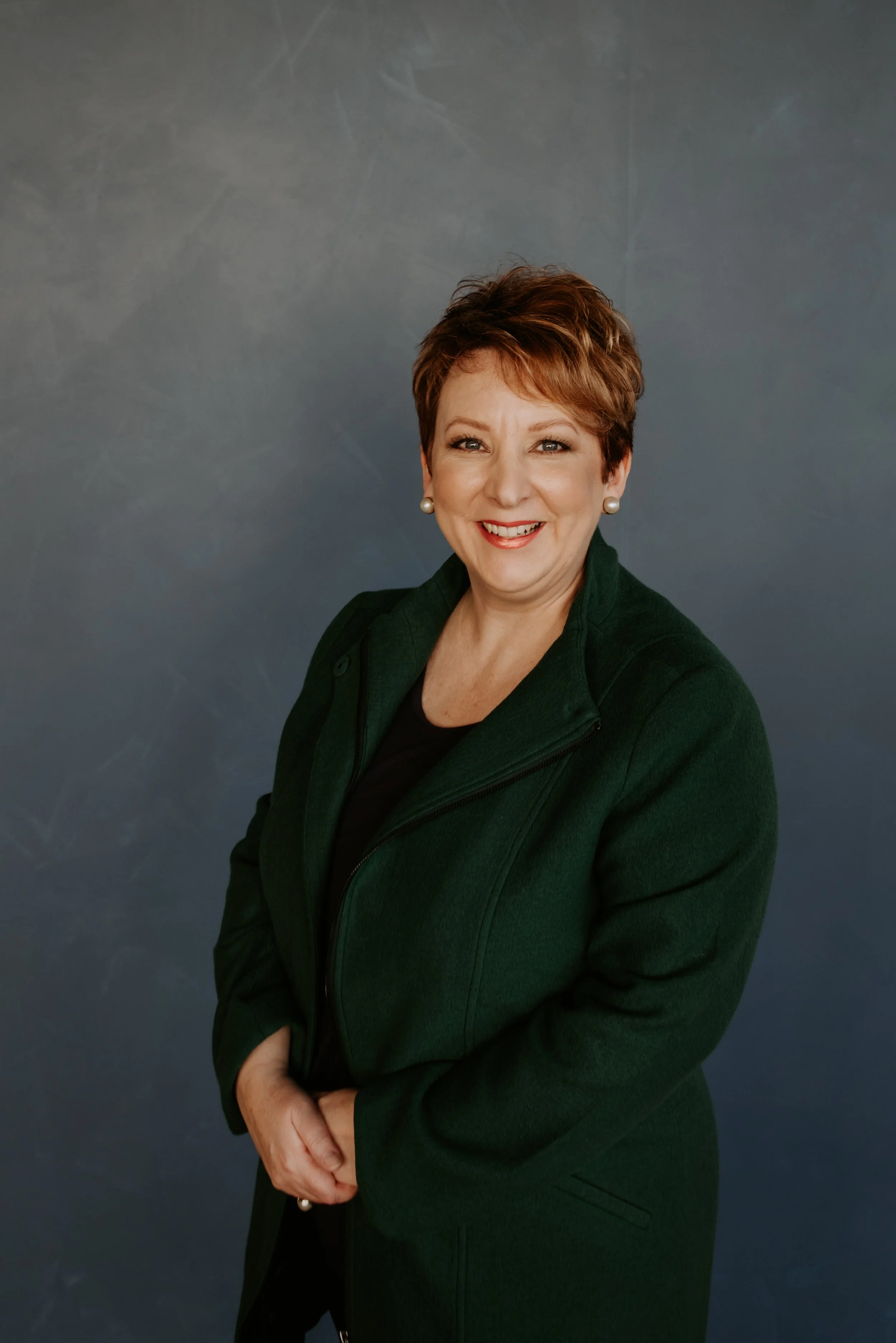 Photo of Emma smiling at the camera. She has short red hair, and wears a dark blazer over a dark shirt.