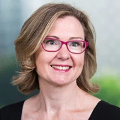 Photo of Trudi smiling at the camera. She has shoulder-length dark blonde hair and wears red-rimmed glasses.
