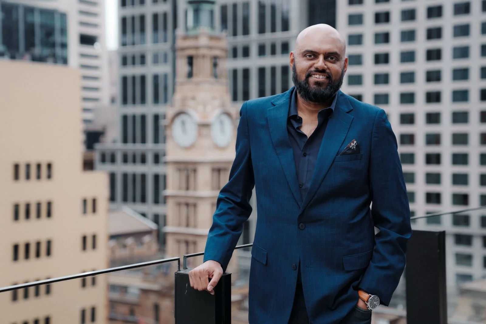 Photo of Bosco smiling at the camera. He has a dark beard and moustache, and wears a navy blazer over a dark collared shirt.