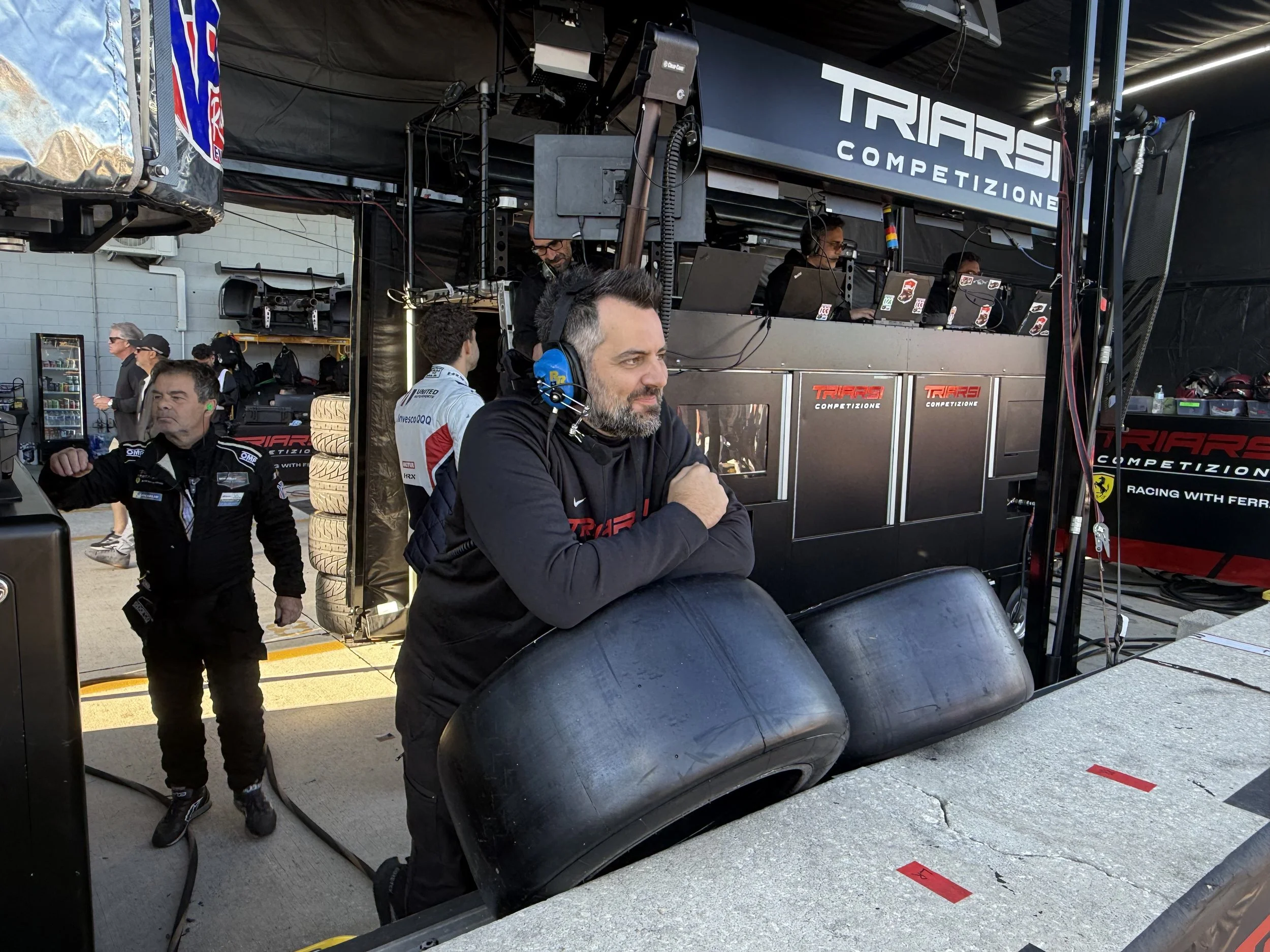 Man in black racing team hoodie leaning on tires with arms crossed, observing racing activity at a garage with people and equipment in the background.