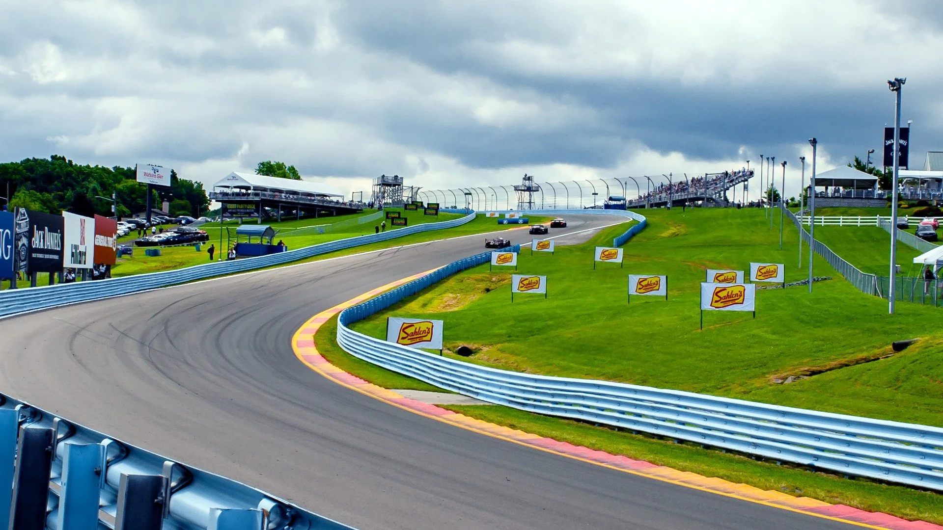 Race track with four cars speeding on a curved section, surrounded by green grass and blue barriers, under cloudy skies, with spectators and banners along the track.