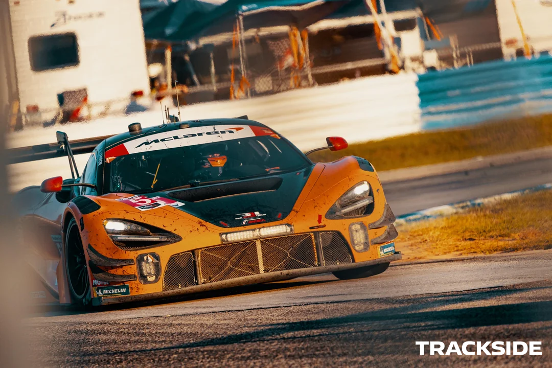 A yellow and black McLaren race car on a racetrack, drifting around a corner during a race, with a blurred background of race structures and a blue sky.