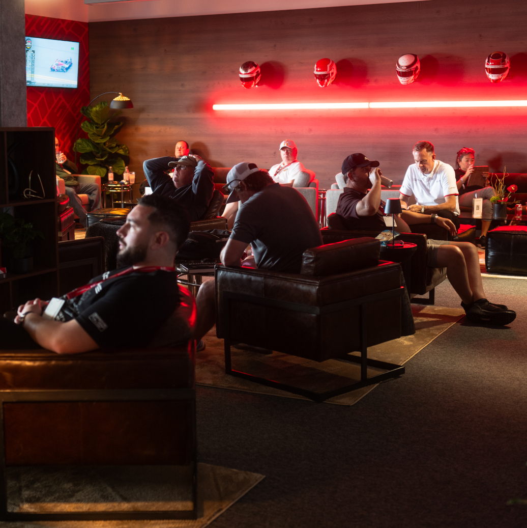 A group of people in a dimly lit lounge area with red lighting, some wearing caps and casual clothing, sitting on black and red chairs, with a wooden wall, helmets on display, and a television screen in the background.