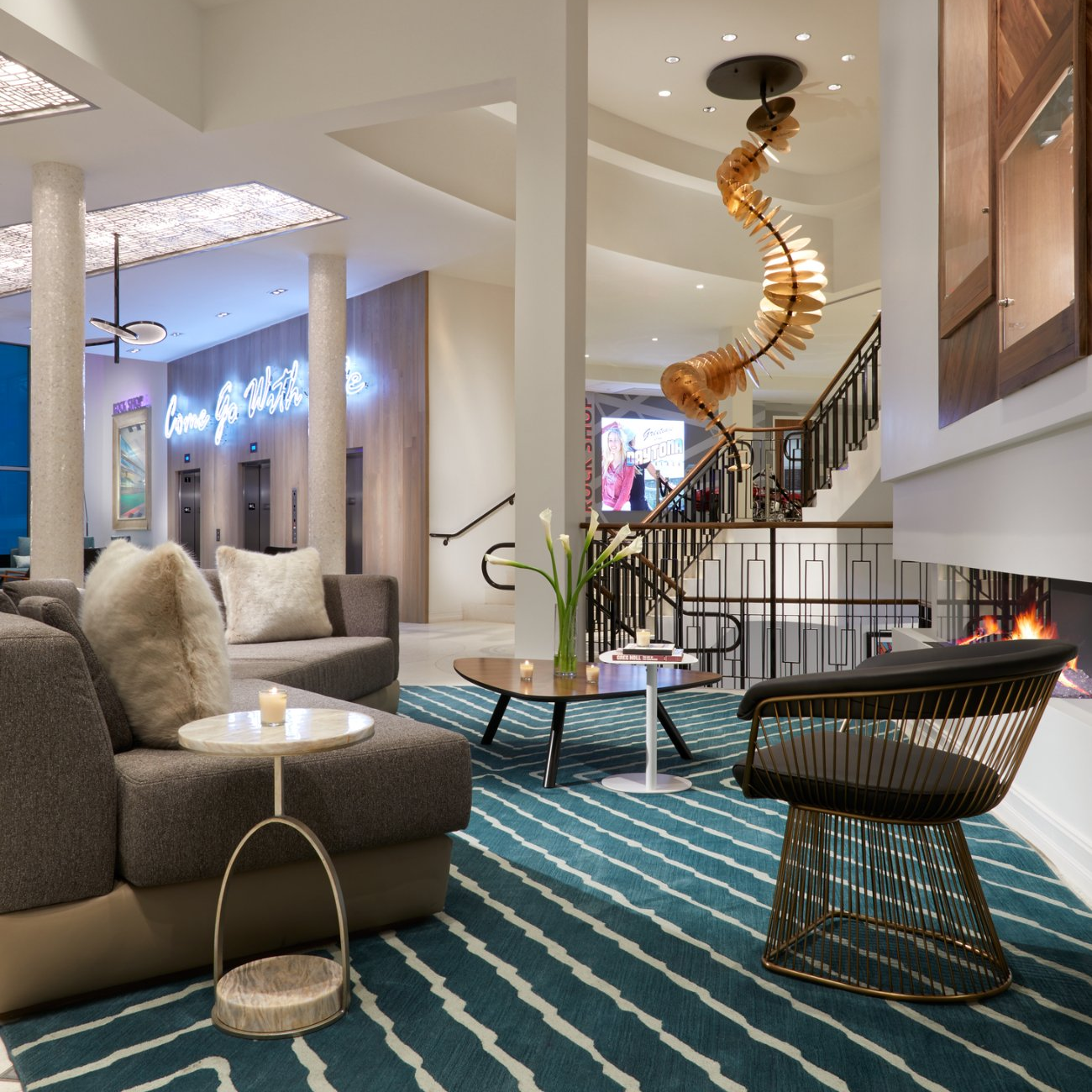 Modern hotel lobby with a gray sofa, fur pillows, a small round side table with a candle, a coffee table with a vase of white calla lilies, a black lounge chair with gold wire frame, a fireplace, a blue and white striped rug, and a spiral wooden sculpture hanging from the ceiling next to stairs.