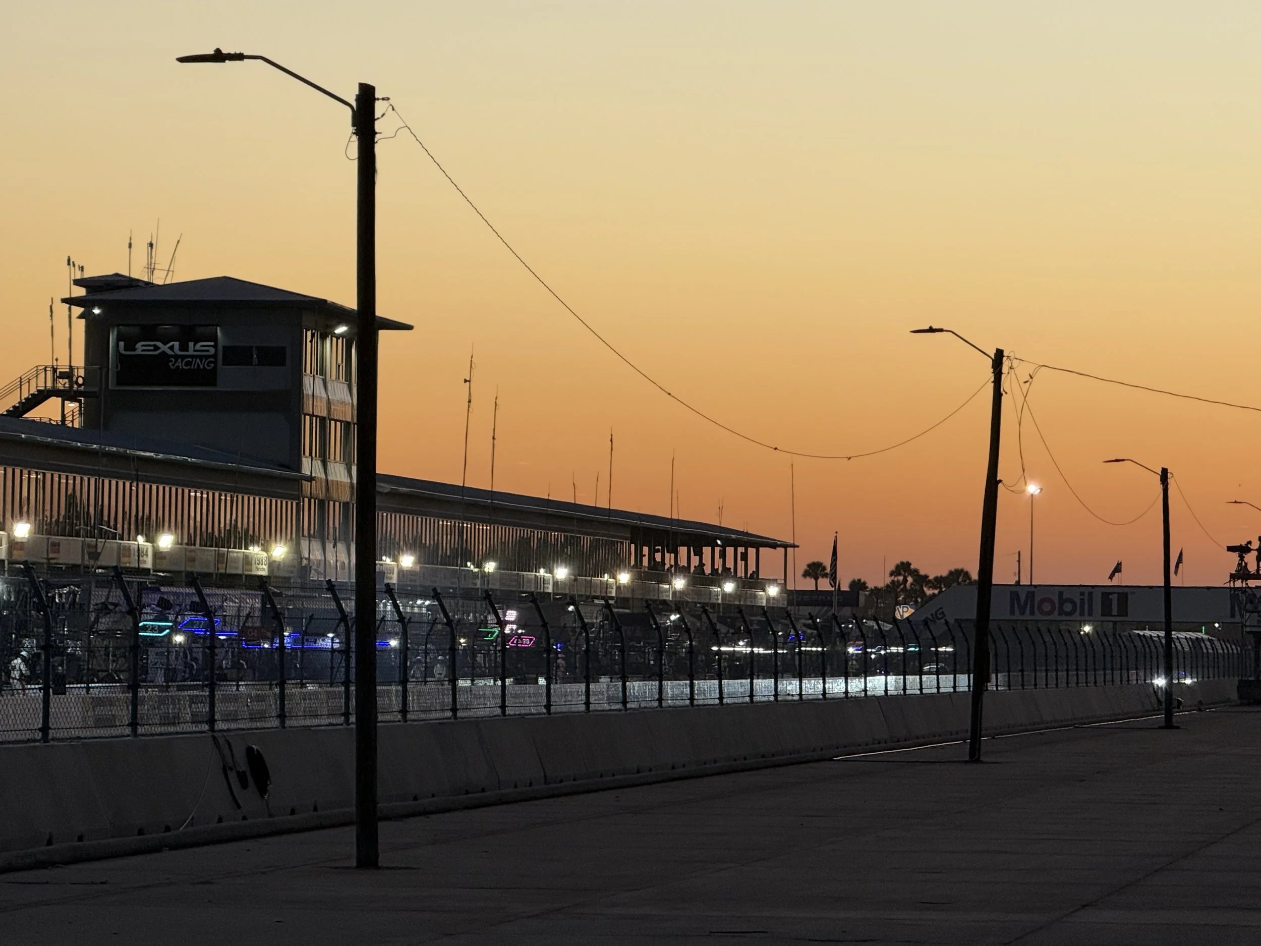 Race track at sunset with lighting, a grandstand with a Lexus Racing sign, and a fence in the foreground.