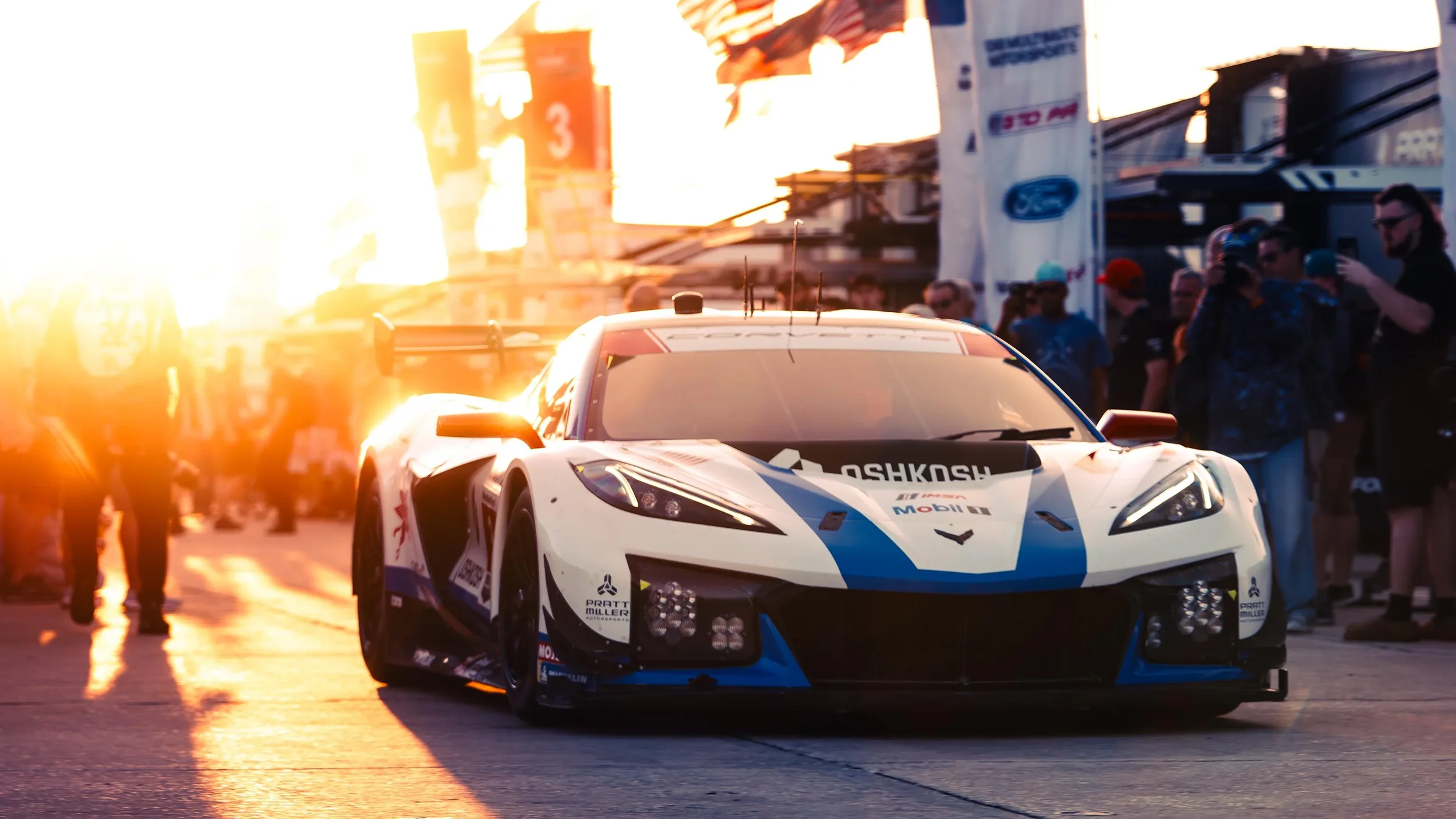 A white and blue race car on a track with people and structures in the background during sunset.