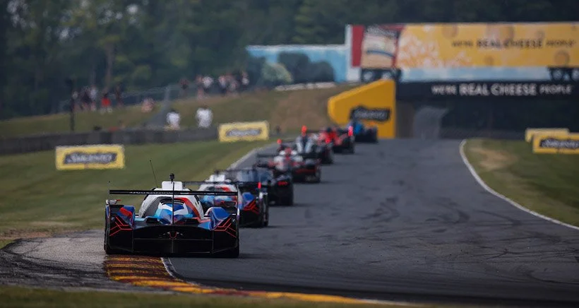 Series of race cars on a racetrack, approaching a turn with a backdrop of trees and advertising banners.
