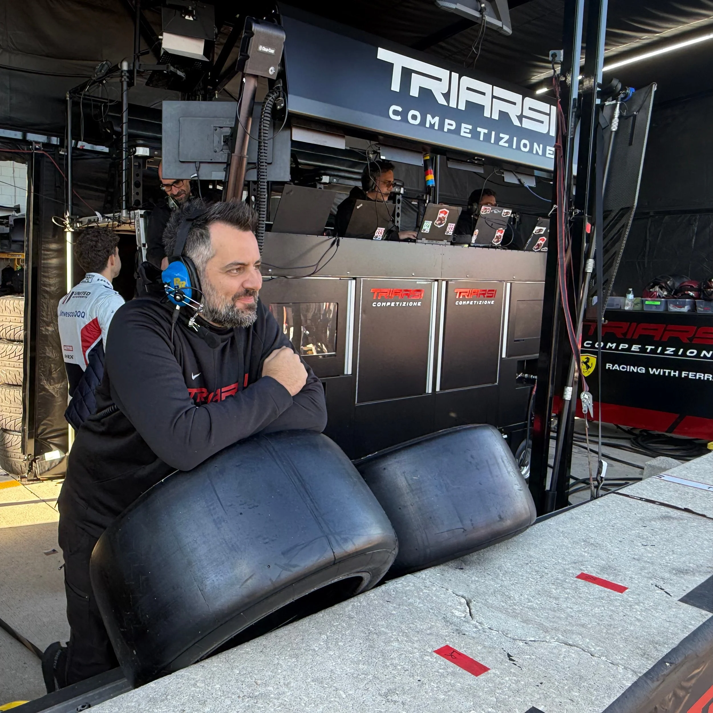 A man with a beard and black hoodie leaning on steered tires in a racing pit, wearing a blue headsets, with a racing team in the background working at their station with laptops and equipment, and a large sign above that reads 'TRIARS COMPETIZIONE'.