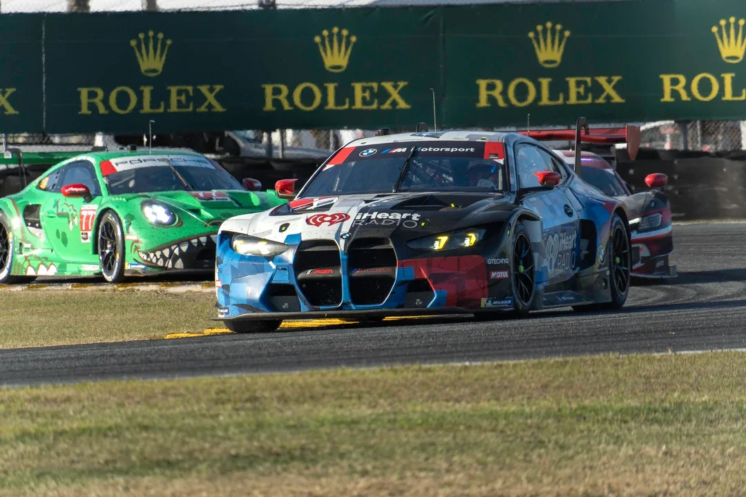 Race car on a track with a green and black banner advertising Rolex in the background, and other race cars following behind.