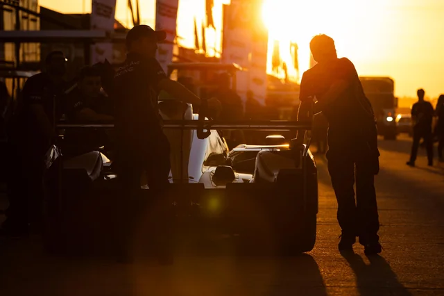 Two individuals standing beside a race car, silhouetted against a setting sun, at a racing event or pit stop.