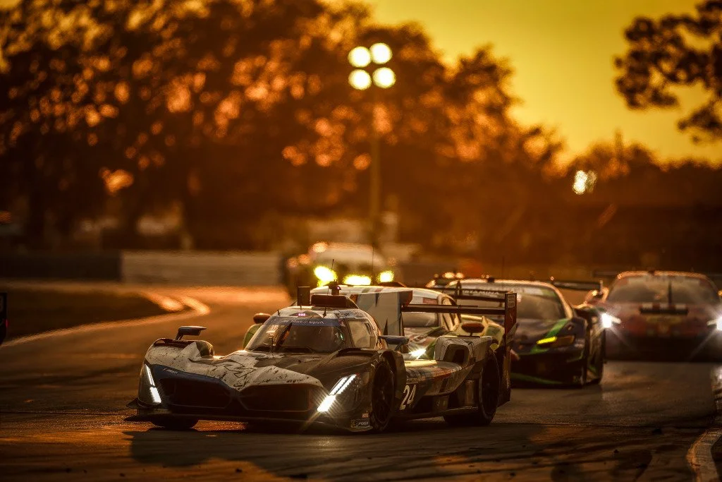 Race cars on a track during sunset, with trees and track lights in the background.