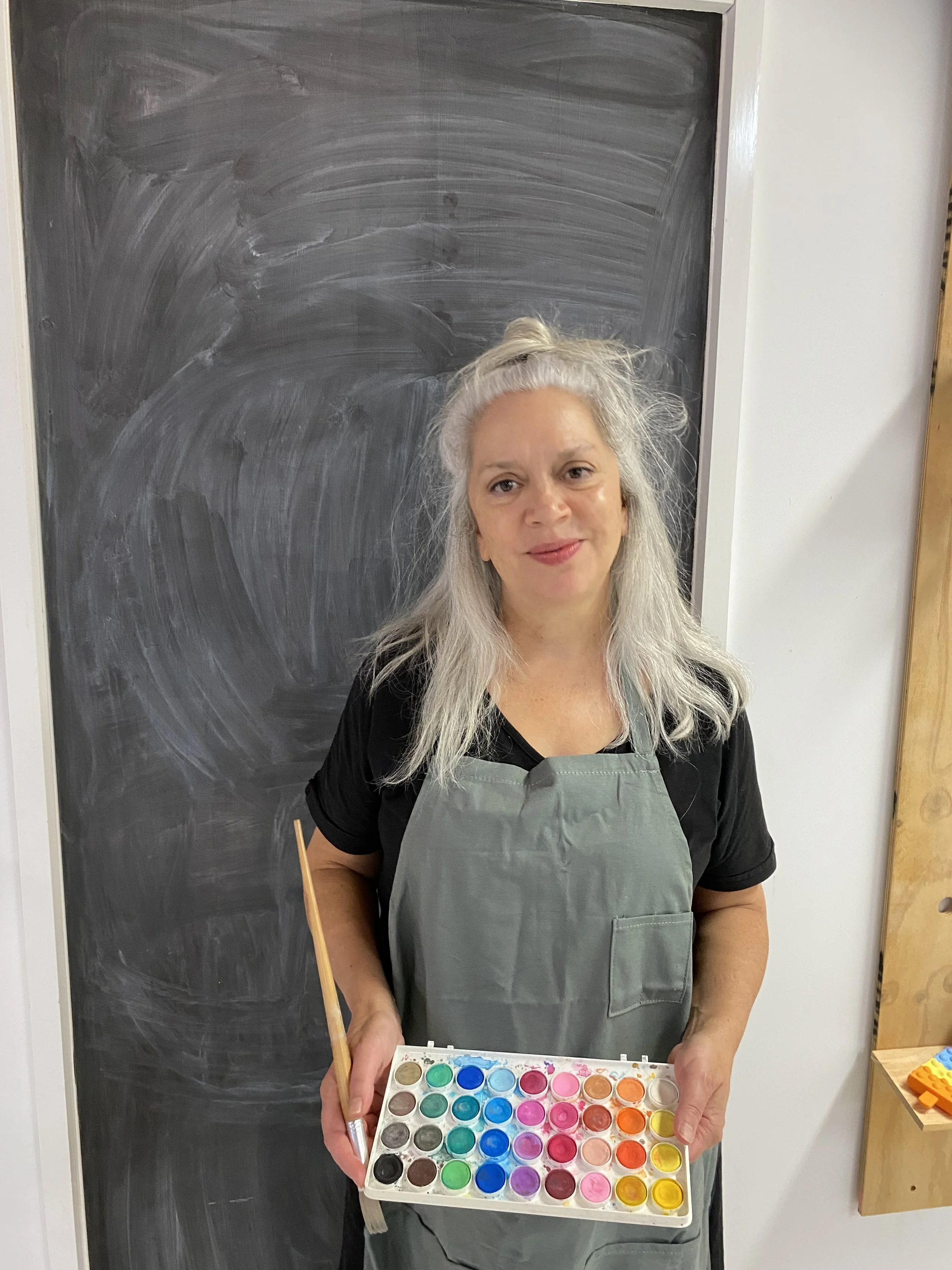 A woman with long gray hair standing in front of a blackboard, holding a palette of watercolor paints and a paintbrush, wearing an apron.