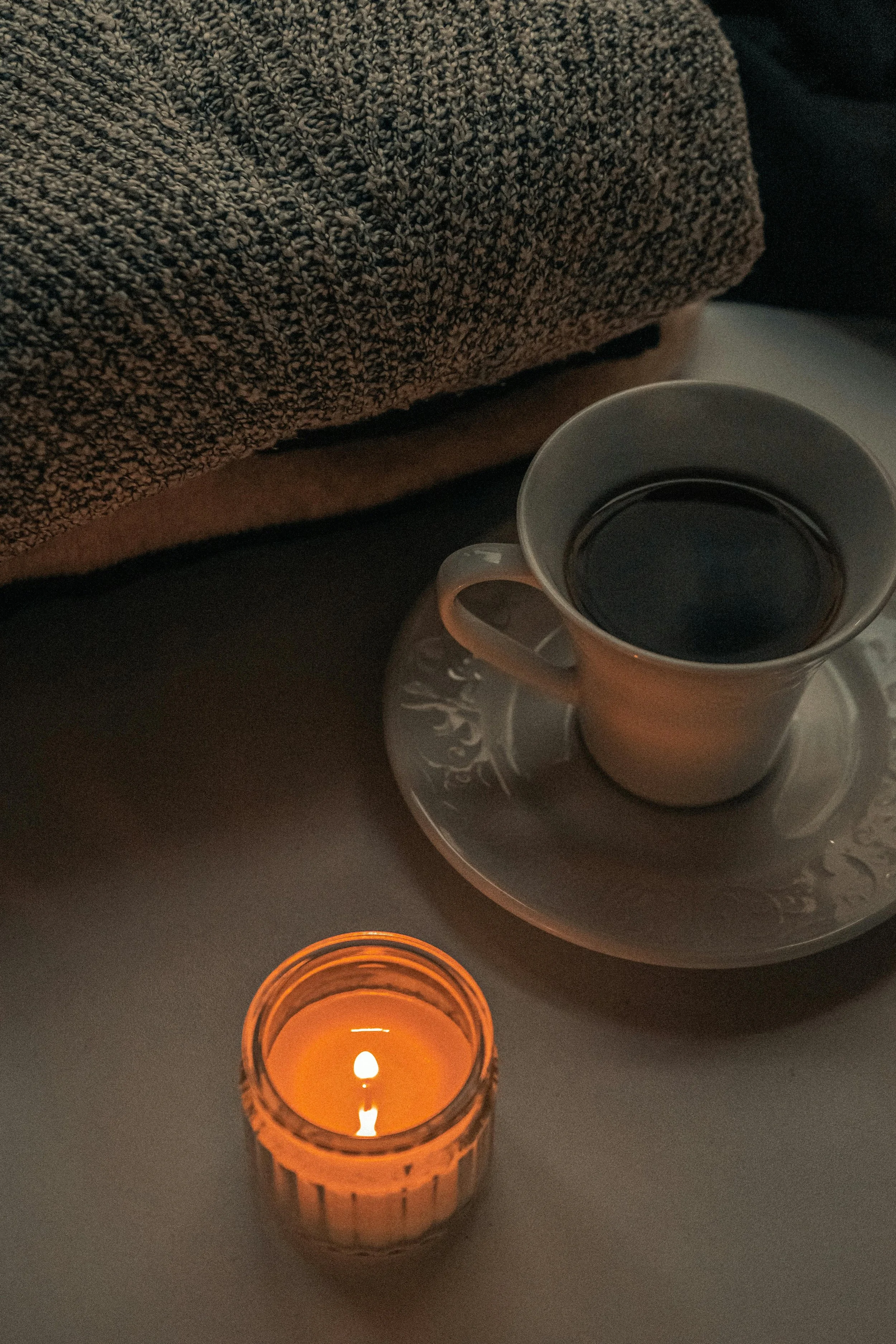 Cozy evening self-care scene with a lit candle, cup of tea, and folded knit blanket on a table in soft warm lighting.