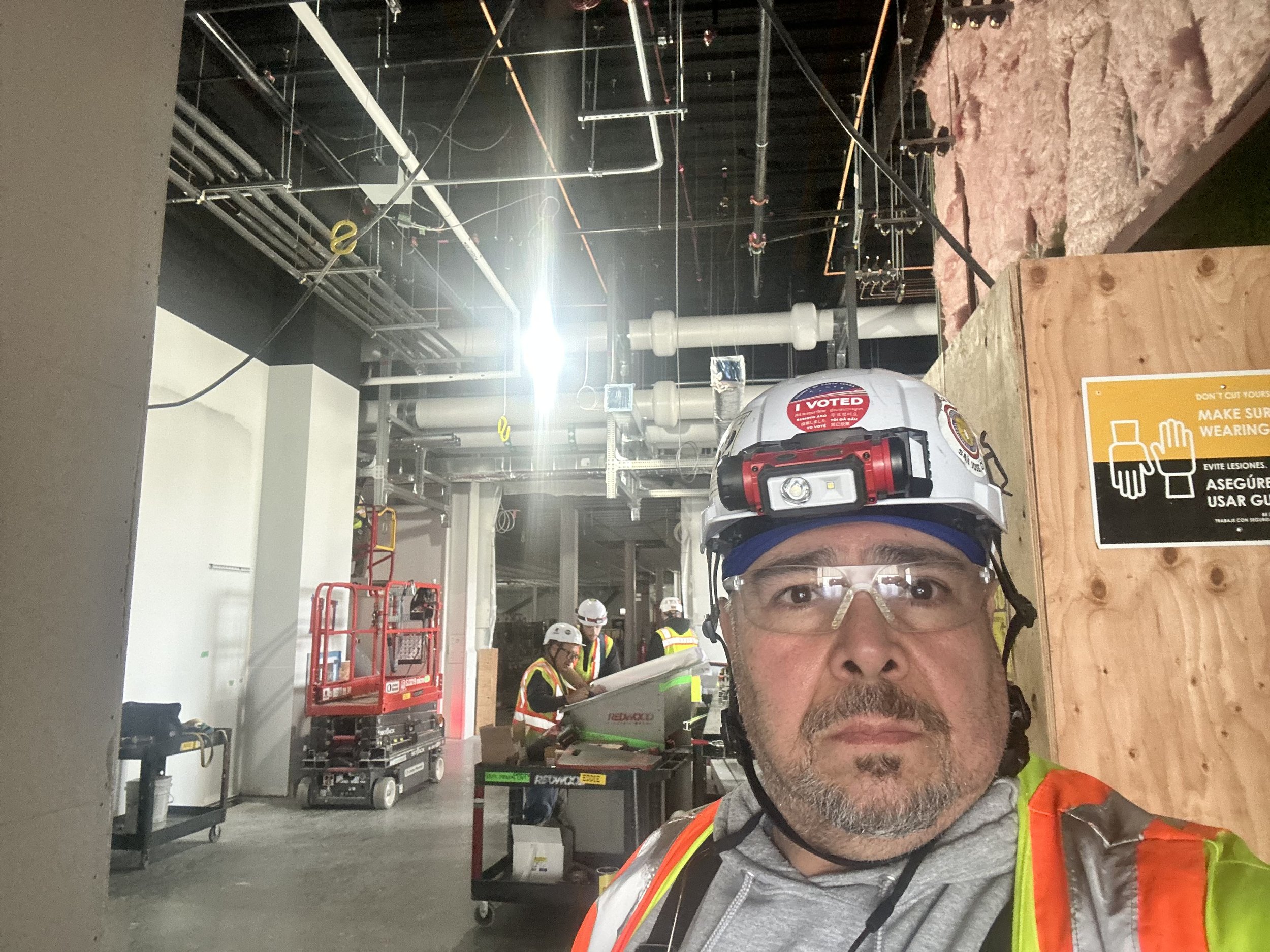 Rafael taking a selfie on a construction site with pipes and workers working in the background.