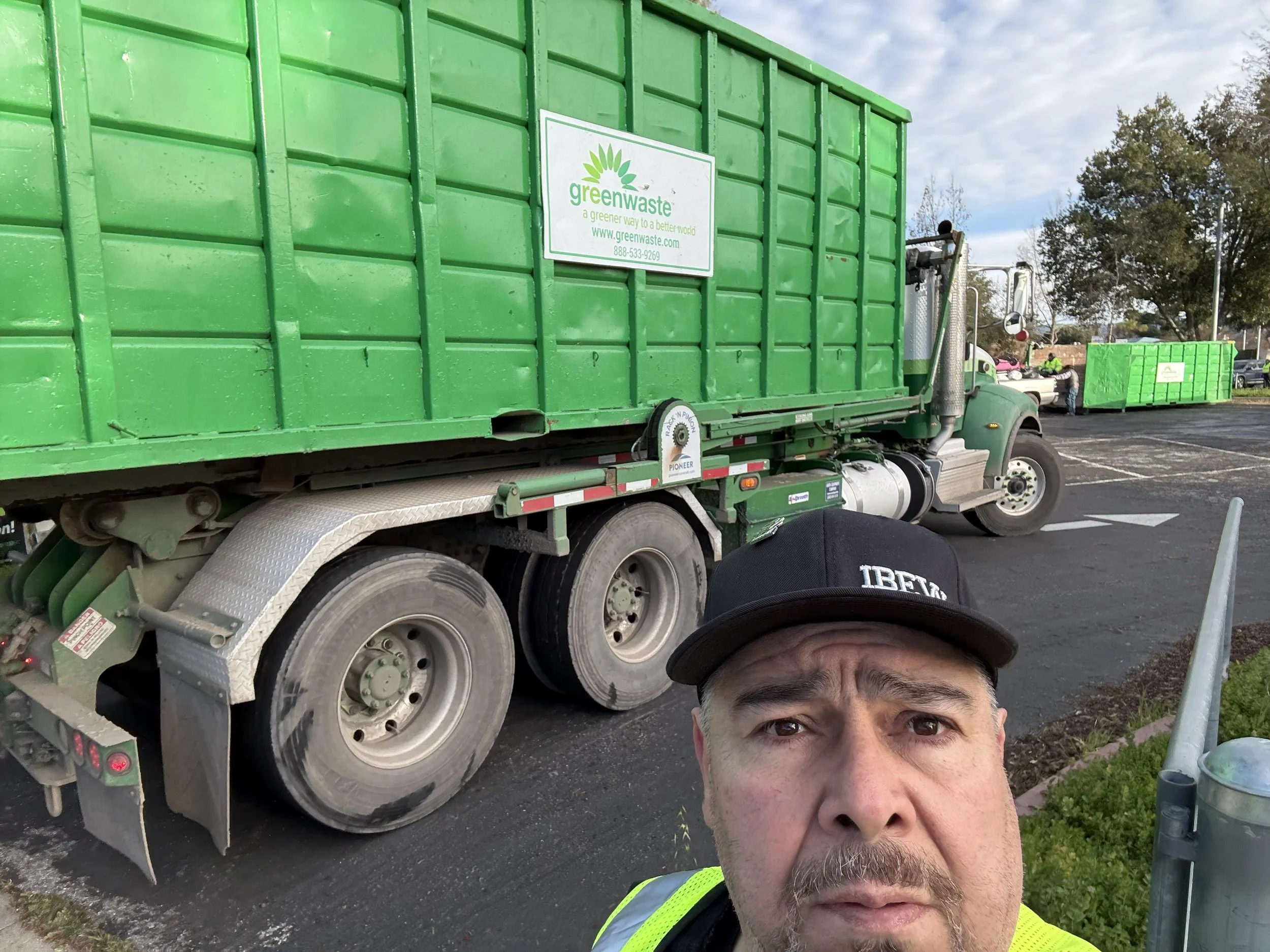 A man taking a selfie in front of a green waste truck in a parking lot. The truck has a green container with a white and green logo that reads 'greenwaste' and a website URL. There is another green waste container in the background and a person standing nearby.