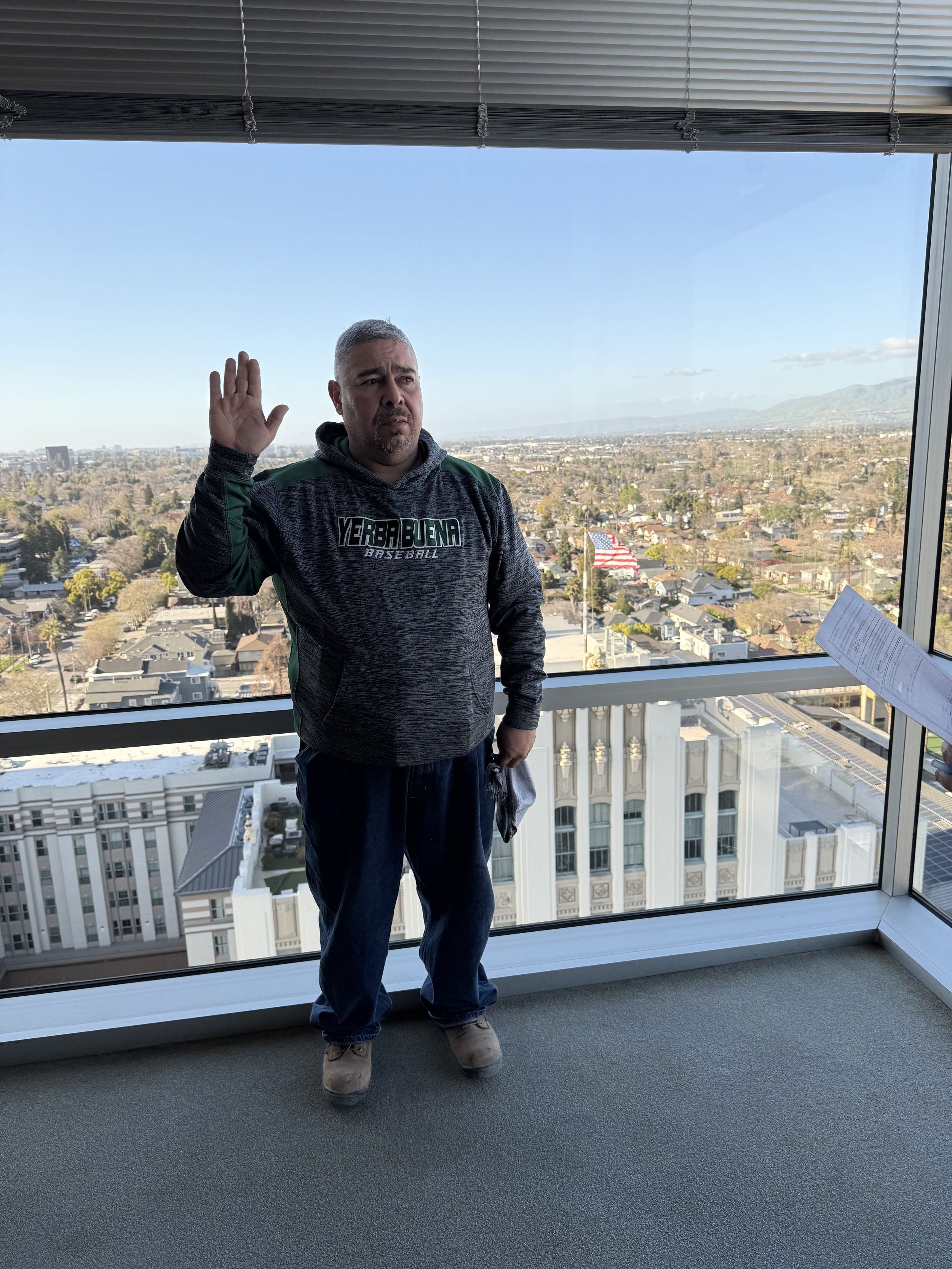 A man standing indoors near a large window with an urban cityscape view, raising his right hand in a greeting or oath, wearing a dark hoodie with 'YERBABUENA BASEBALL' logo, jeans, and work boots.