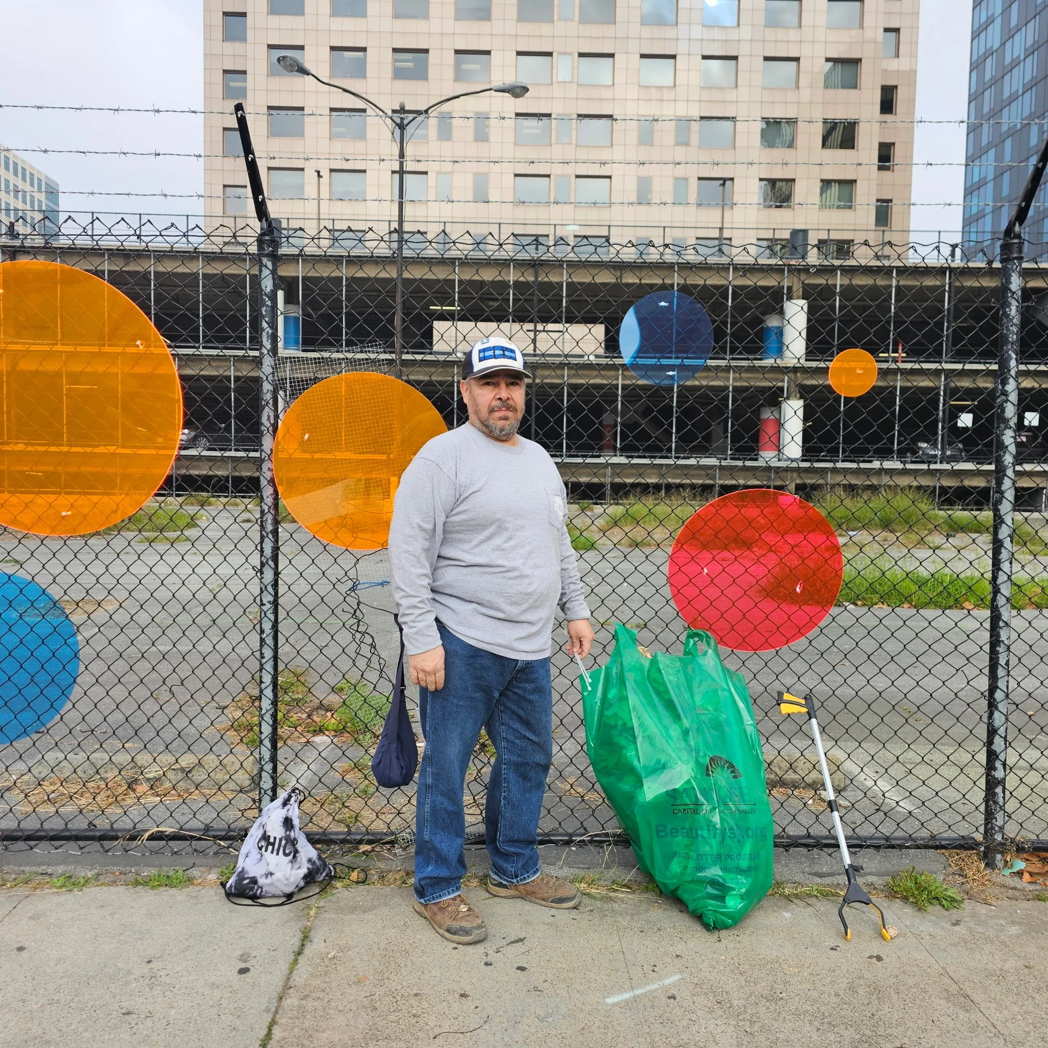 Rafael standing in front of a chain-link fence decorated with colorful circles, holding a large green trash bag and a green trash grabber, with a city building behind him.