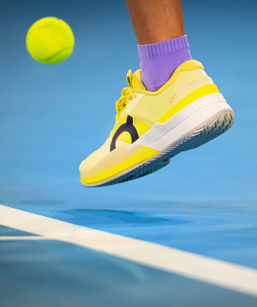 A yellow and white tennis shoe with a black logo on a person's foot, wearing a purple sock, about to hit a tennis ball on a blue tennis court.
