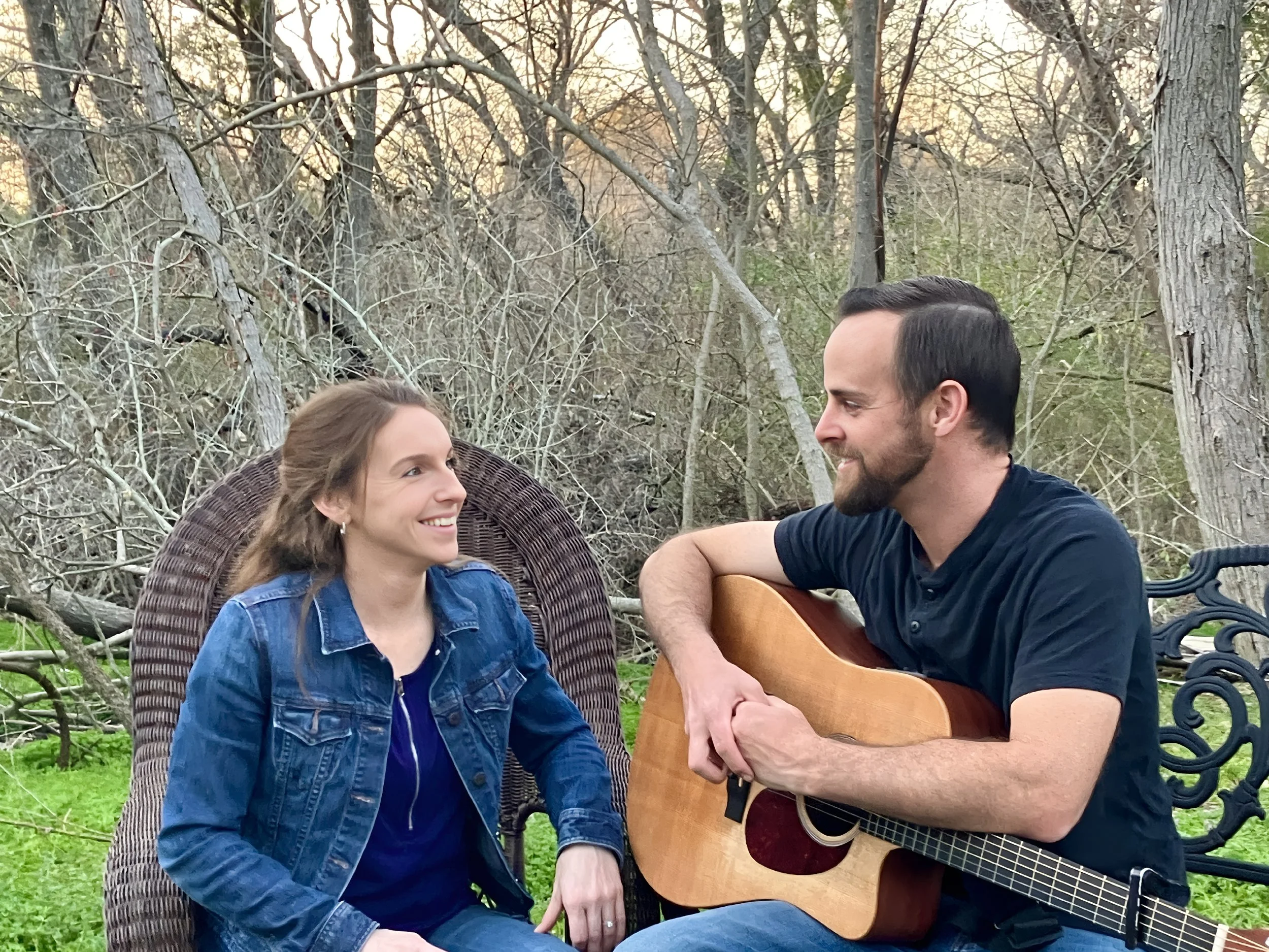 A man and woman sitting outdoors in a wooded area, with the man playing a guitar and the woman smiling at him.