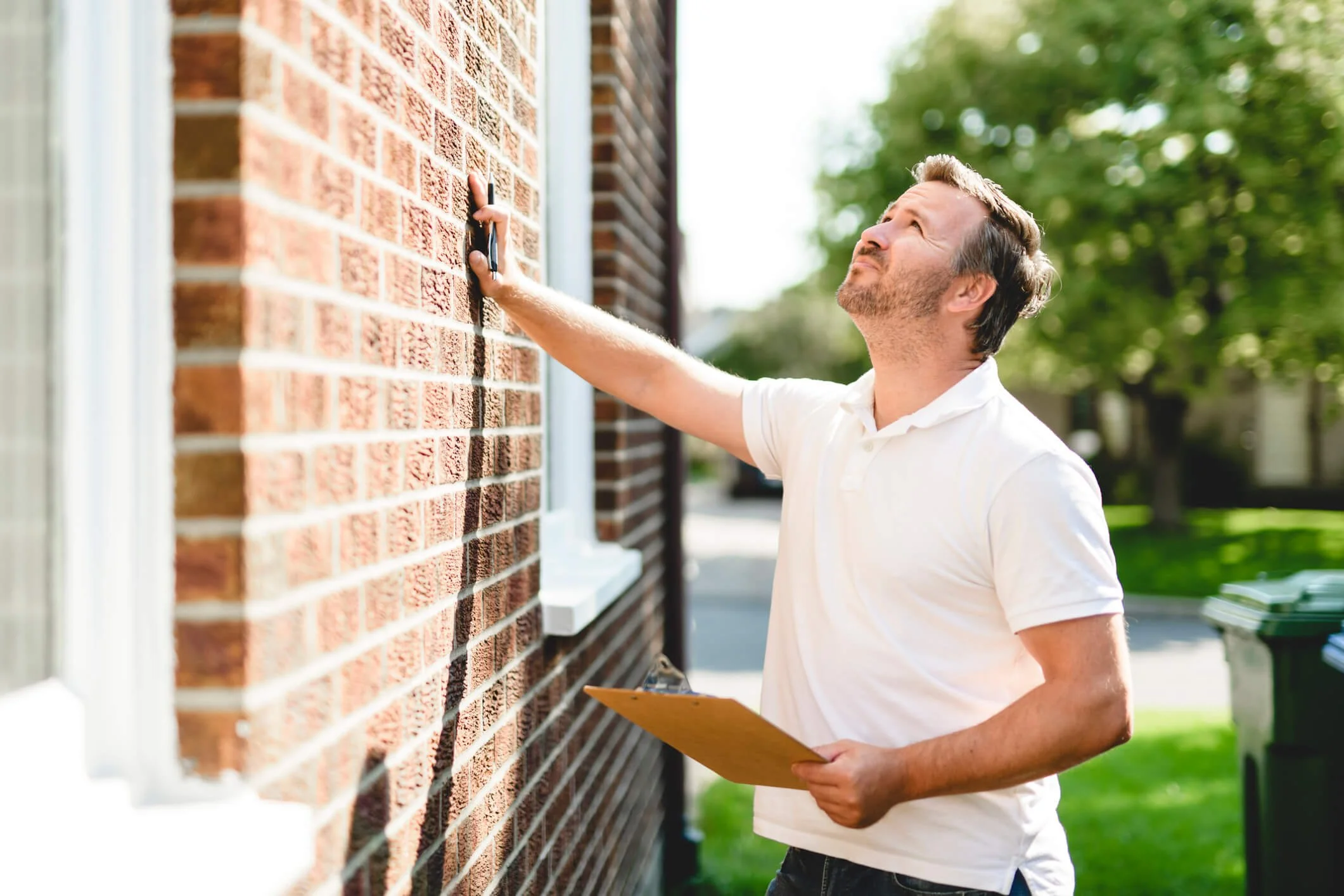 Man with a clipboard inspecting or marking a brick wall outside.