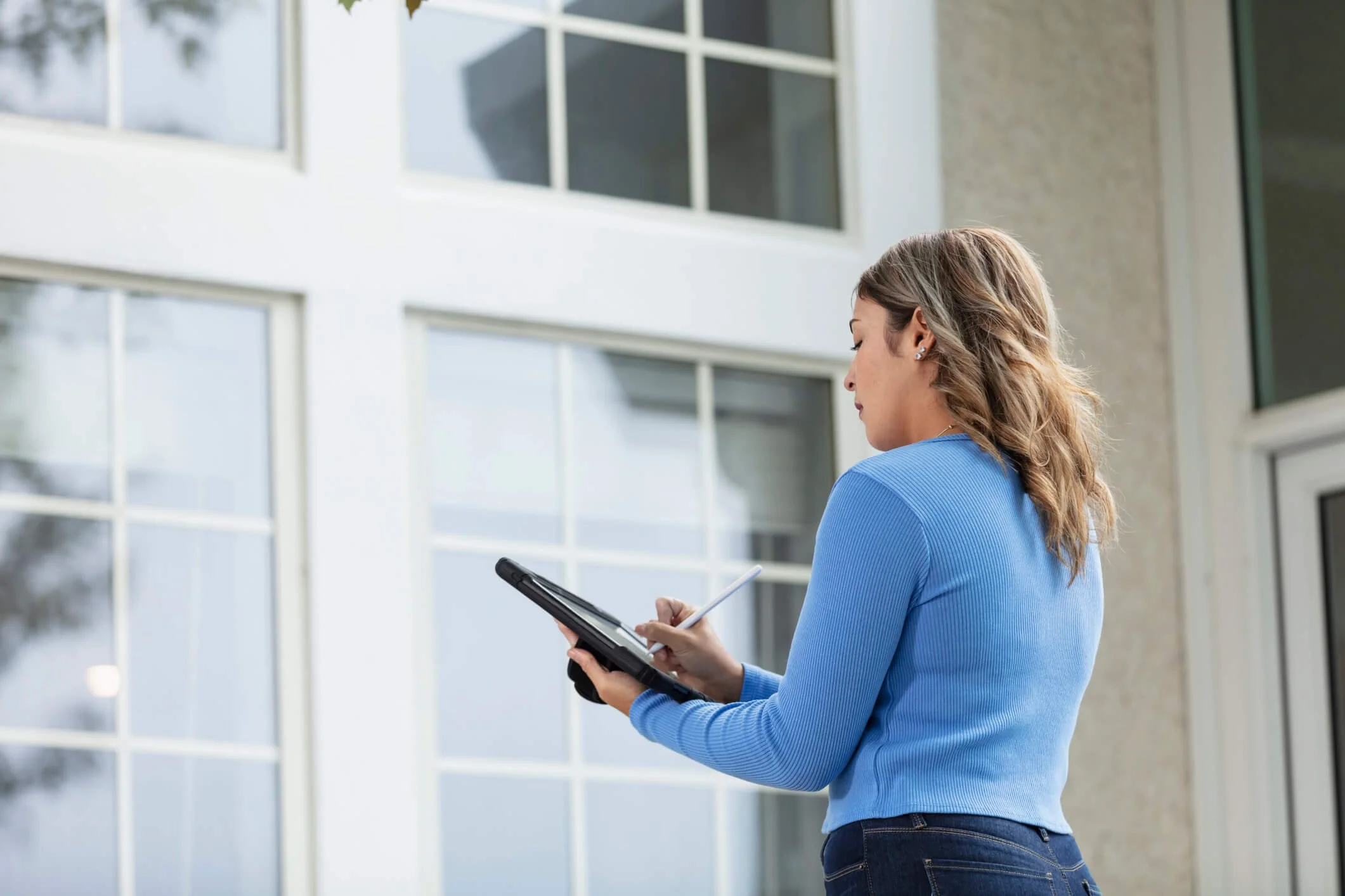 Woman in blue sweater standing outside in front of a modern building, holding a tablet and a stylus, looking at the device.