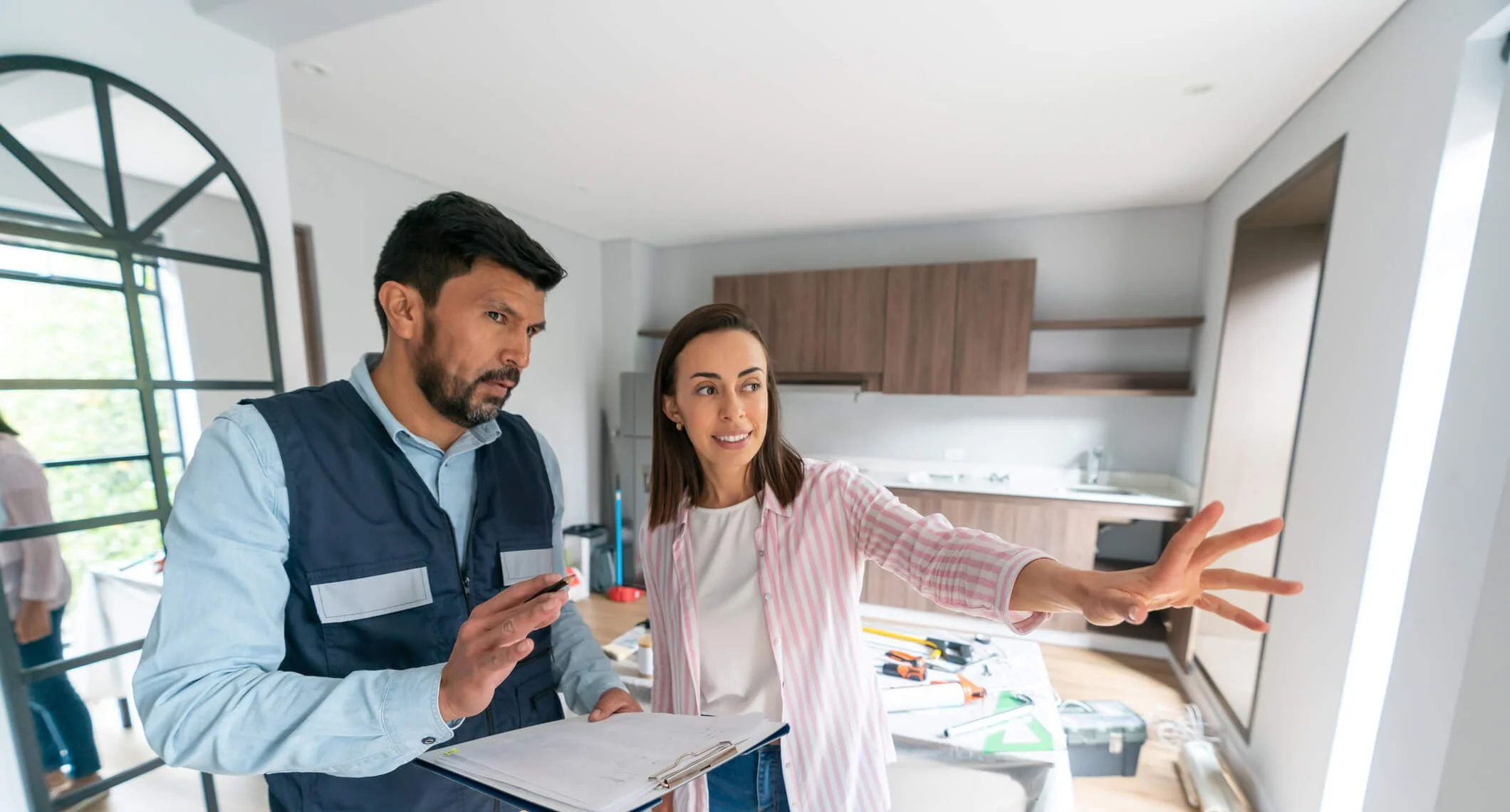 A man in a blue vest and a woman in a pink striped shirt discussing renovation plans in a modern kitchen, with tools and papers on the counter.