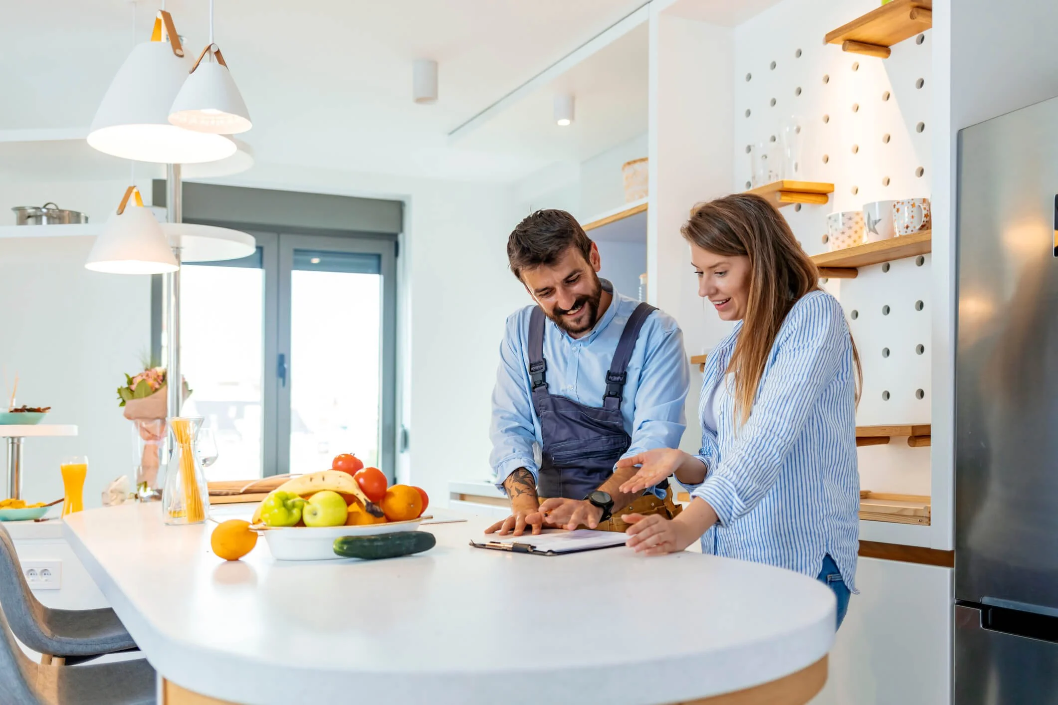 Two people, a man and a woman, are happily looking at a notebook on a kitchen island with fresh fruits and vegetables.