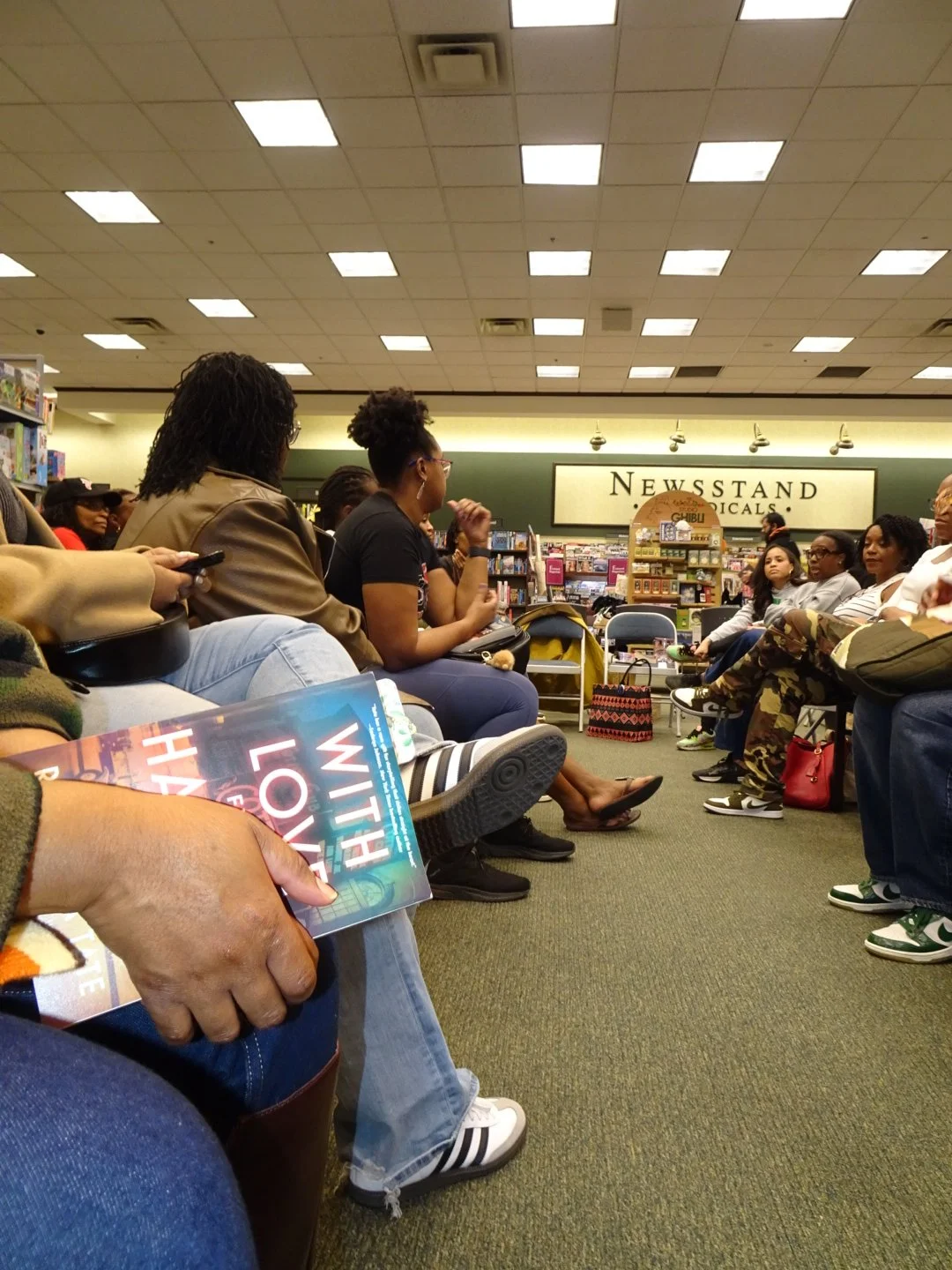 Group of people seated in a bookstore, participating in an event or book signing. Shelves and a sign reading "NEWSSTAND" are visible in the background.