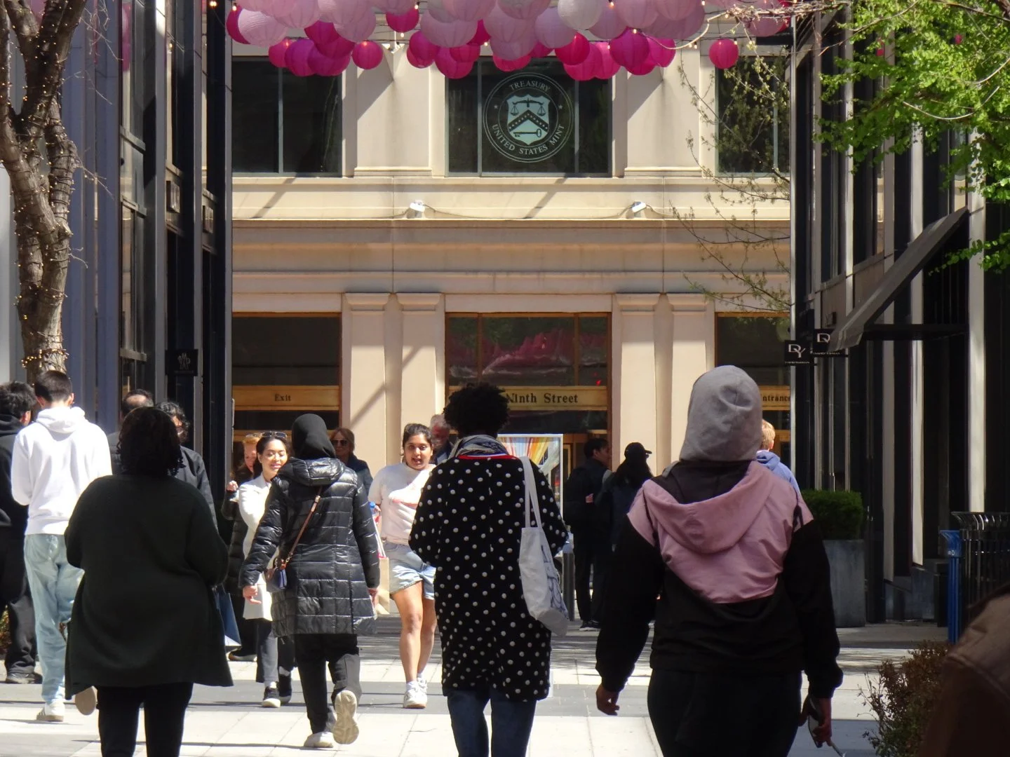 People walking through a busy outdoor shopping area with storefronts, trees, and decorative pink and purple paper lanterns overhead.