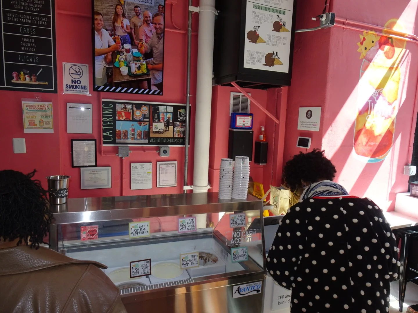 Inside a pink ice cream shop with a counter displaying various ice cream flavors. Two people are visible, one wearing a black and white polka dot jacket and the other in a brown leather jacket. The wall features framed menus, posters, and a televisio