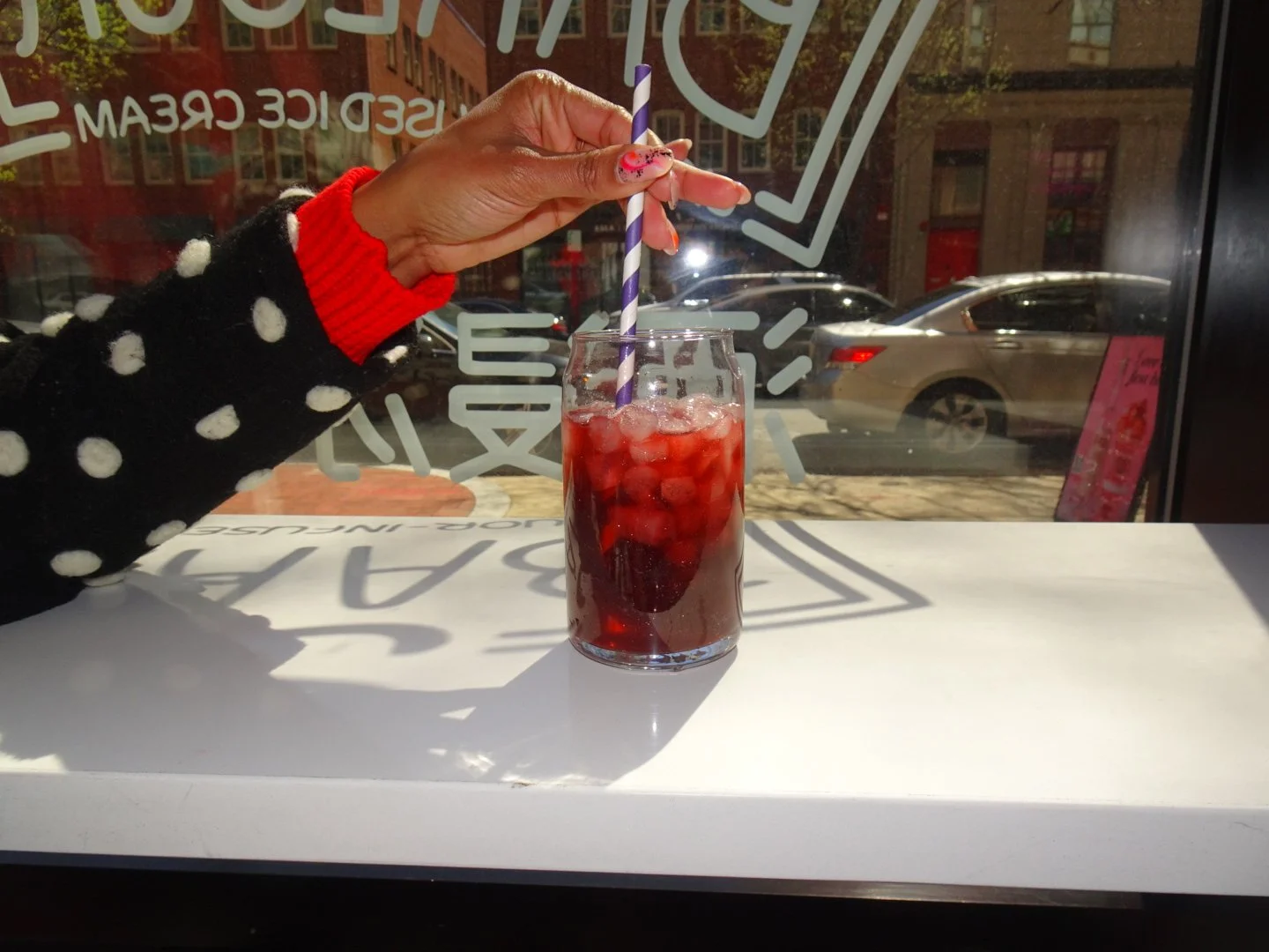 A person with a red sleeve and black polka dot coat holding a purple and white striped straw in a glass of cherry soda with ice, sitting by a window with cars parked outside.
