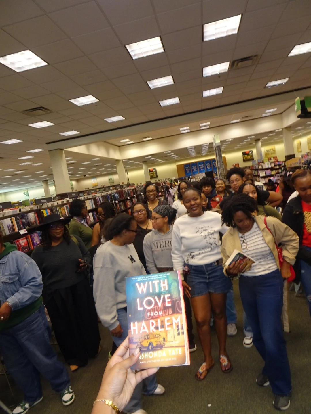 A person holds a copy of the book "With Love from Harlem" in front of a group of women inside a bookstore.