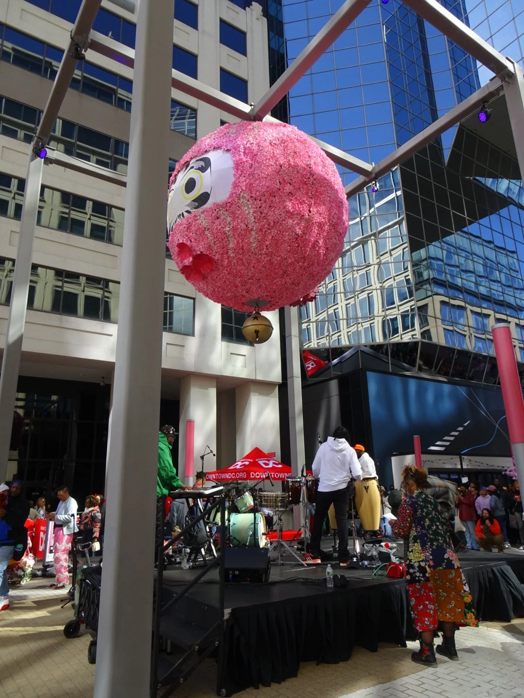 Large pink spherical lantern with a Daruma doll face design, hanging above an outdoor stage with musicians and a crowd, in an urban setting with glass buildings.