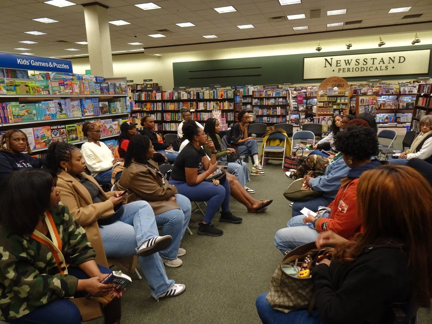 A group of people sitting in a circle inside a bookstore or library, engaged in a discussion or event. There are shelves of books and games in the background, with a sign that reads 'NEWSSTAND PERIODICALS'.