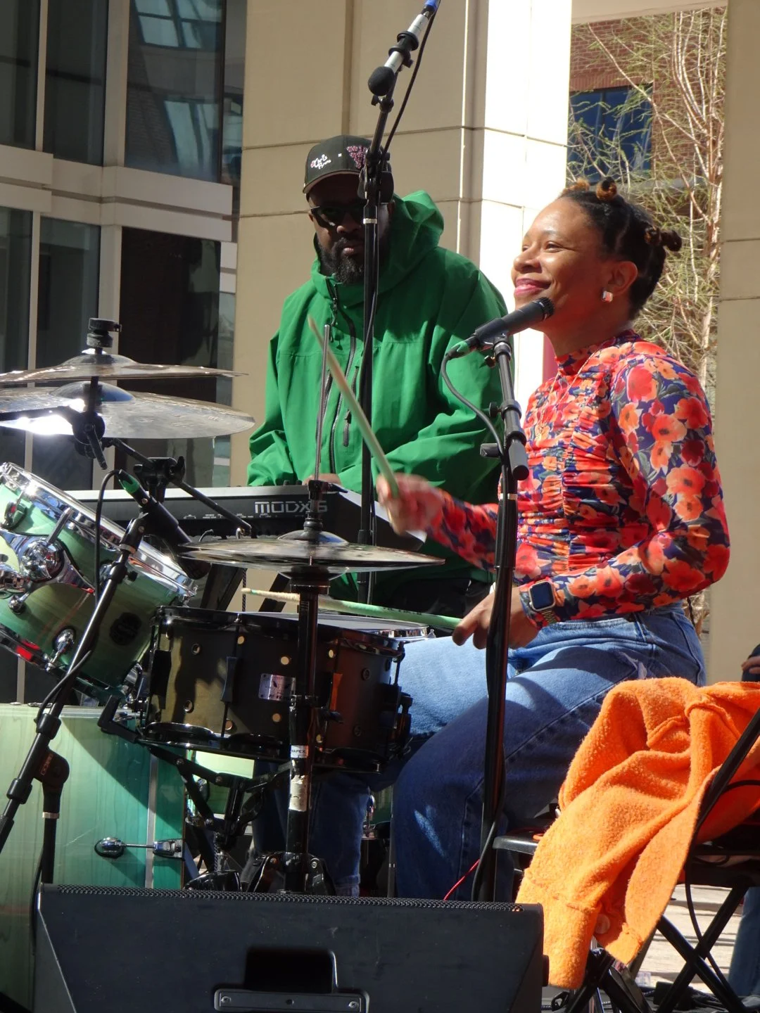 A woman and a man performing music outdoors. The woman is singing into a microphone and playing drums, while the man is playing a keyboard. They are surrounded by buildings and trees.