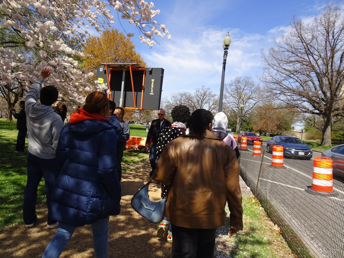 People standing outside in a park during daytime, with some trees blooming, a construction barrier along the roadside, and a large screen or sign being set up.
