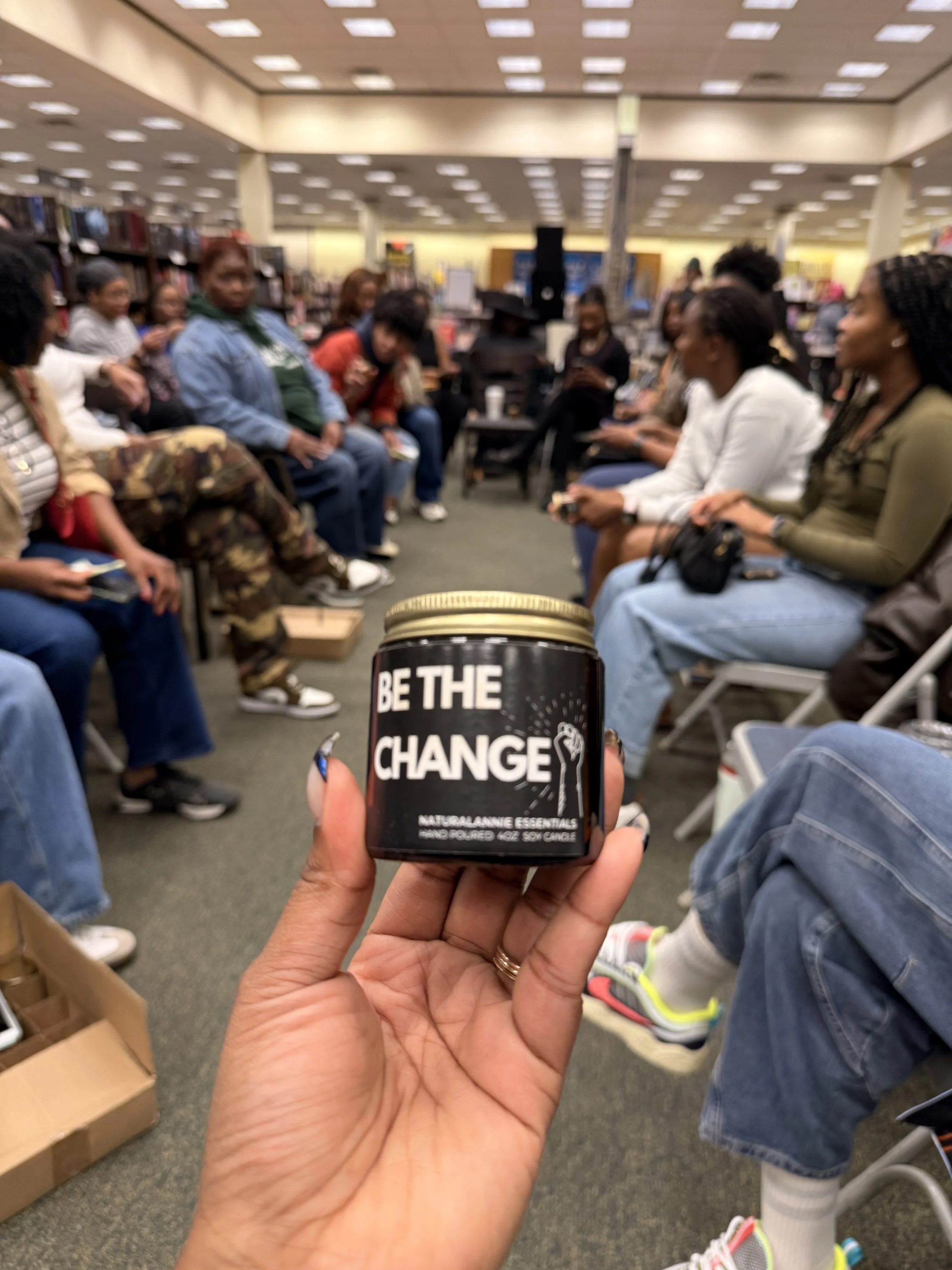 A hand holding a black container with the phrase 'Be the Change' in front of a seated audience in a bookstore or library.