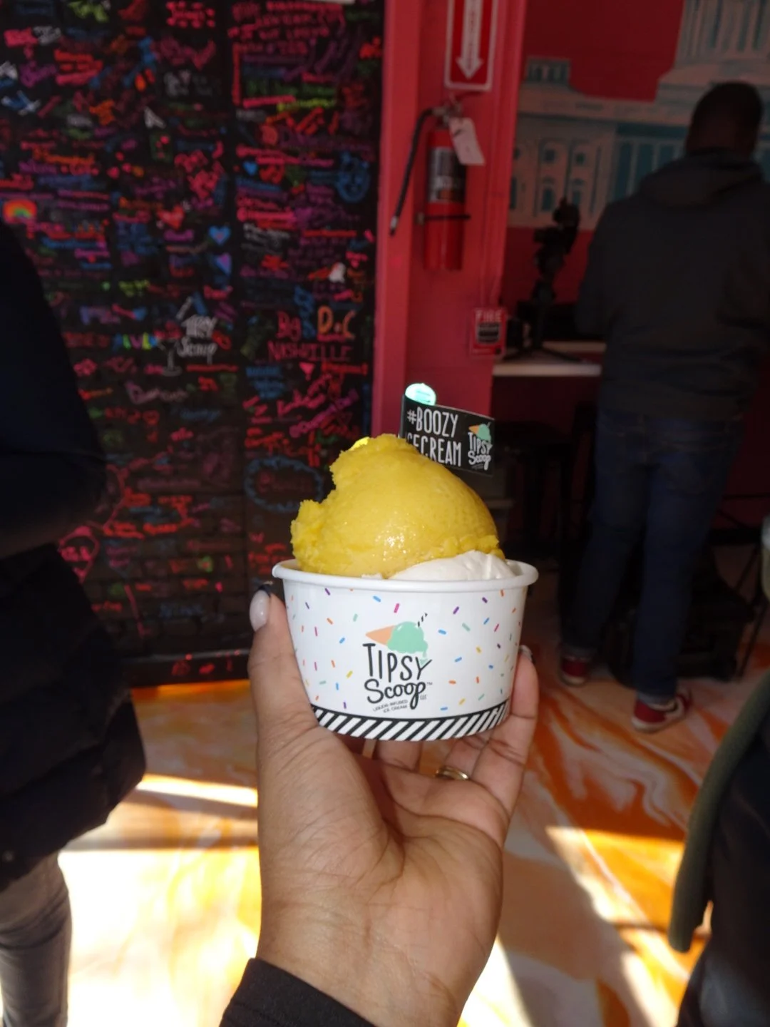 A person holding a cup of yellow ice cream from Tipsy Scoop with a black flag that says "#BOOZY ICE CREAM" inside a colorful, lively indoor establishment.
