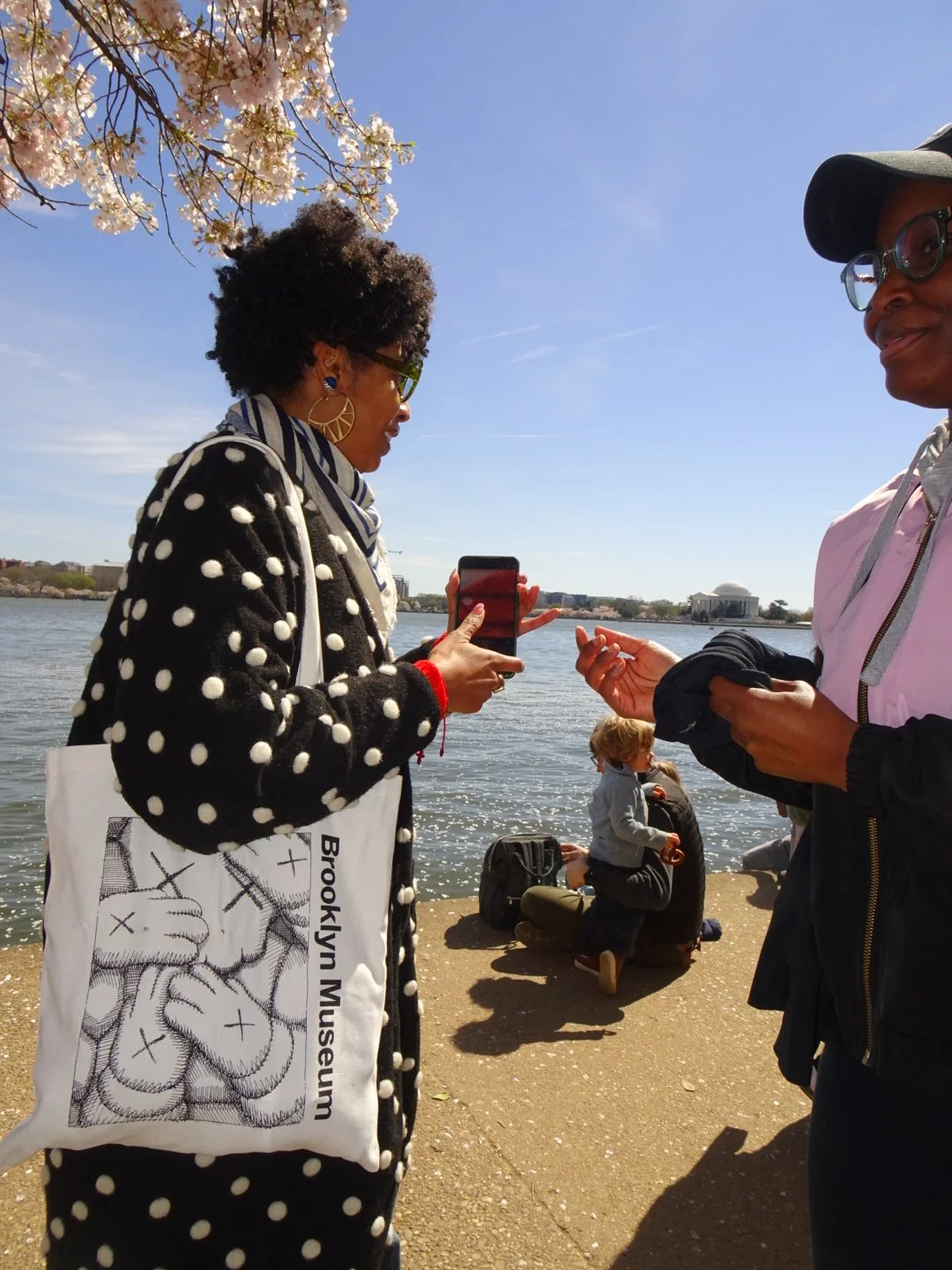 Two women conversing by the riverbank during a sunny day, one taking a photo or video with her phone, with a cherry blossom tree and people, including a child, in the background.