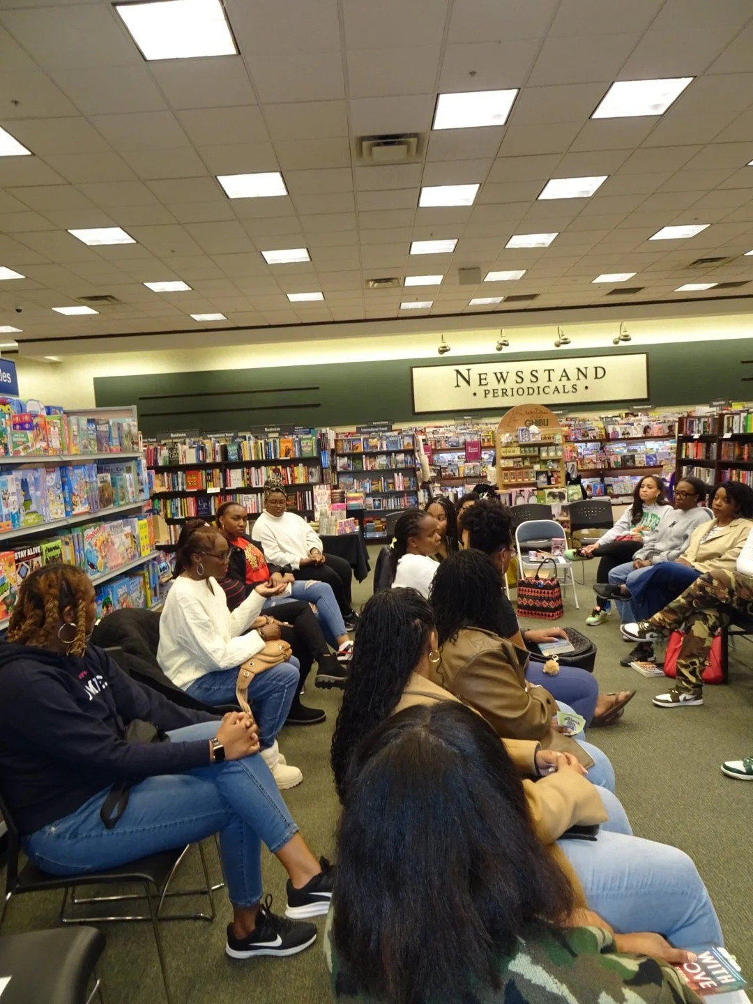 Group of people sitting in chairs in a bookstore, engaged in a discussion or event, with bookshelves and a sign that reads 'Newsstand Periodicals' in the background.