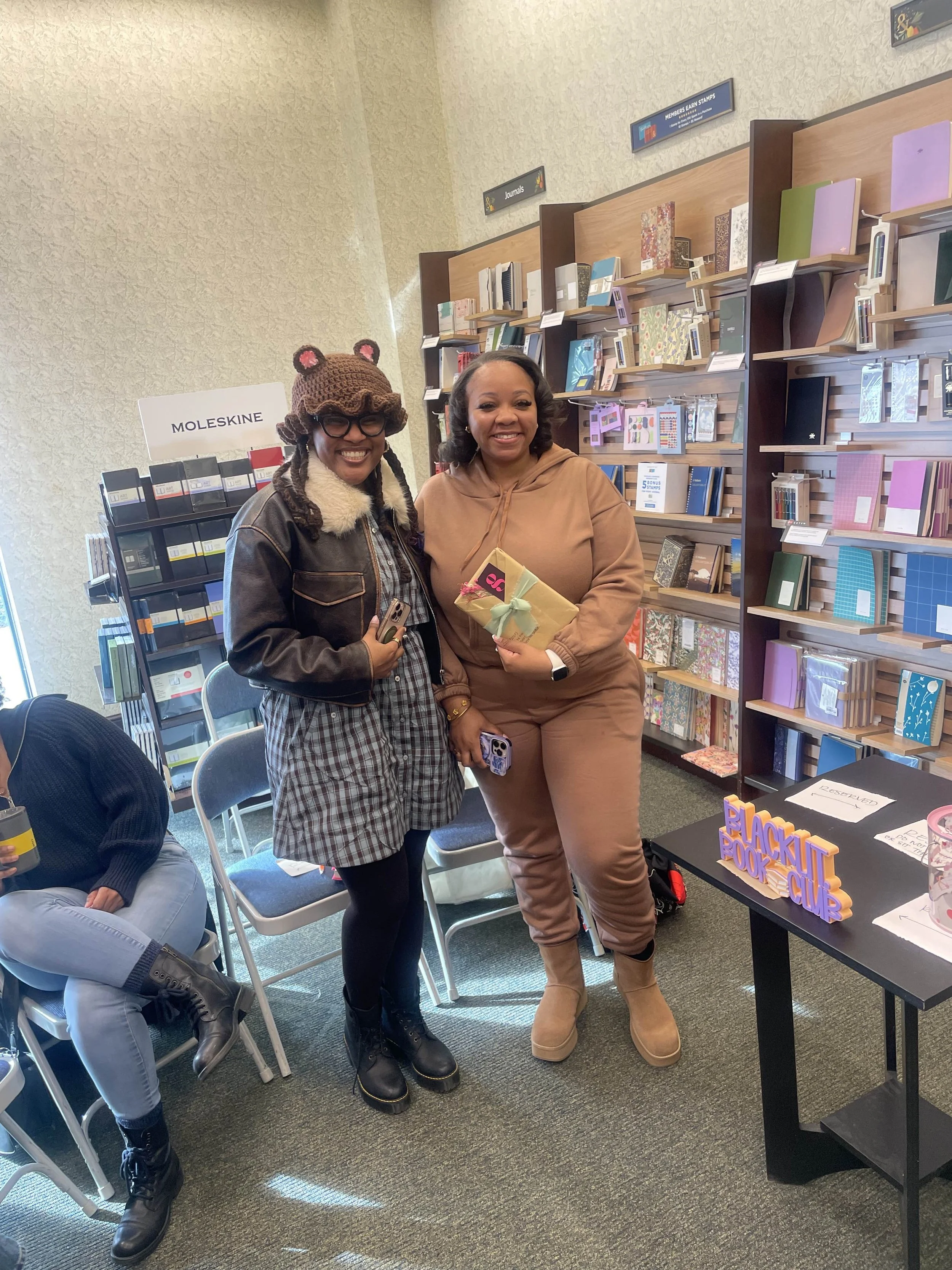 Two women standing in a bookstore, smiling and holding wrapped gifts. One is wearing a bear hat and glasses, the other in a brown hoodie and boots. bookshelves filled with journals and books are behind them.