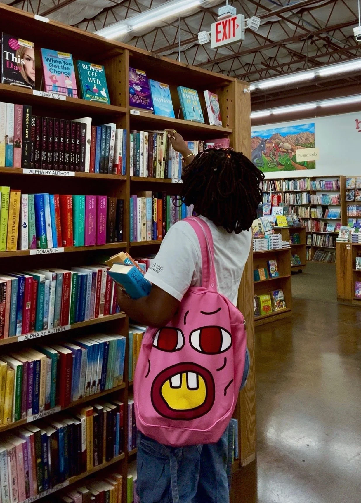 Tye, a woman with locs wearing a white T-shirt and blue jeans shopping for books in a bookstore, carrying a pink otote with a cartoon face, and holding a book in her left hand while selecting books from the shelf with her right hand.