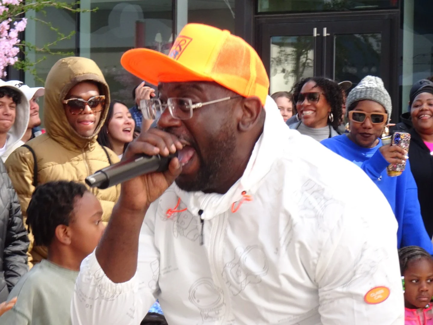 A man wearing glasses and an orange cap singing into a microphone during an outdoor event surrounded by smiling people, including children and women, some wearing sunglasses and hats.