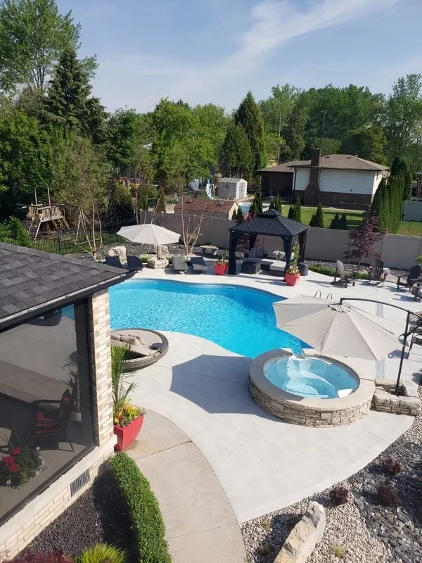 An after swimming pool installation backyard with a hot tub, surrounded by patio furniture, umbrellas, and greenery under a sunny sky.