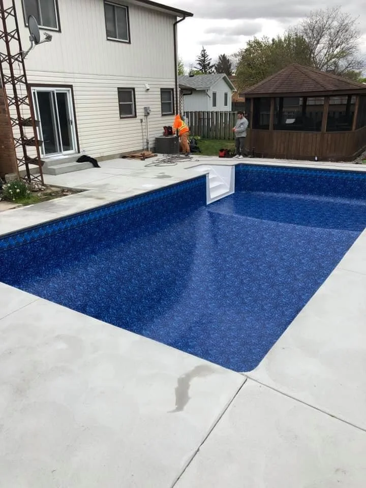 After backyard renovation, with a new in-ground swimming pool with blue liner, surrounded by concrete patio, with two people working near the house and a wooden gazebo in the background.