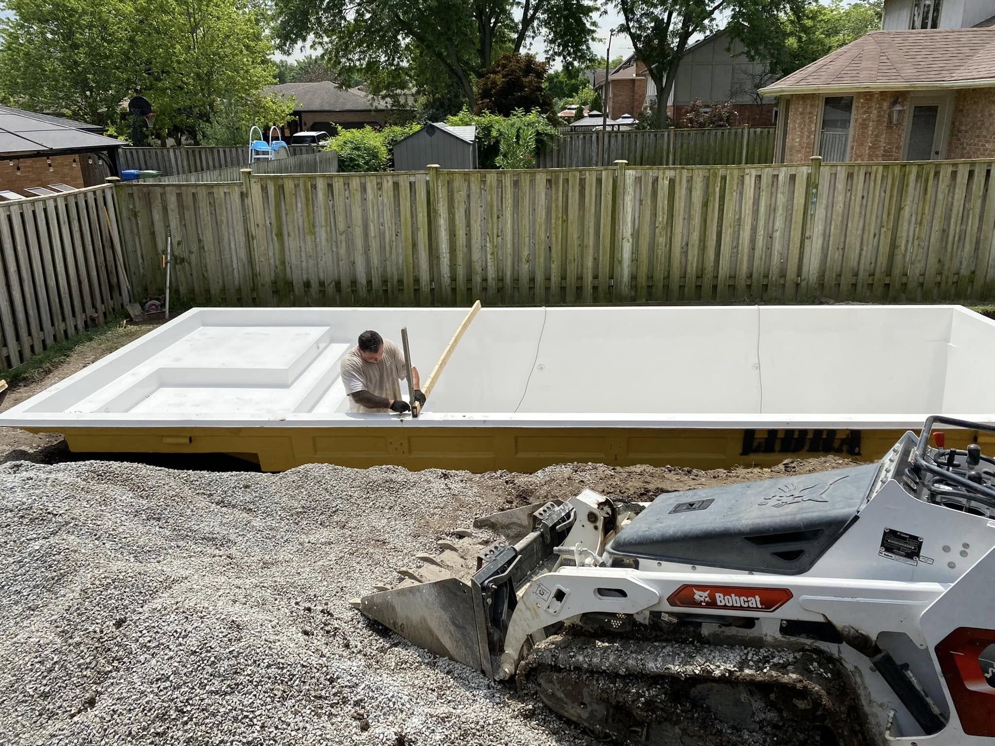 A man building an inground swimming pool in a backyard, with a forklift nearby, surrounded by a wooden fence and residential houses.