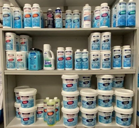 Shelf filled with containers of hot tub chemicals, mostly in white and blue bottles, including spansules and jar sizes, with a blue bottle of a hot tub cleaning or disinfectant product on the middle shelf.
